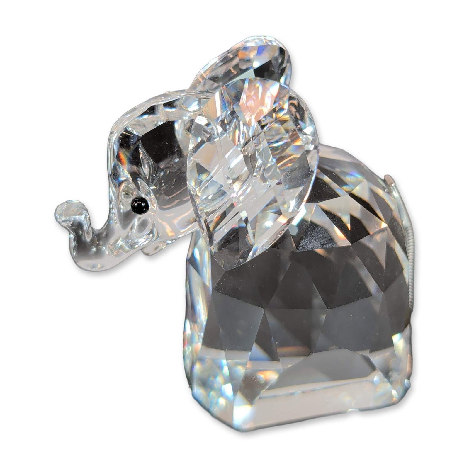 Swarovski Crystal Elephant Figurine: Elephant figurine in clear faceted crystal, featuring a curved lowered trunk and two black crystal bead eyes. The piece is finished with a flexible silver-tone coiled metal wire tail. Swarovski etched