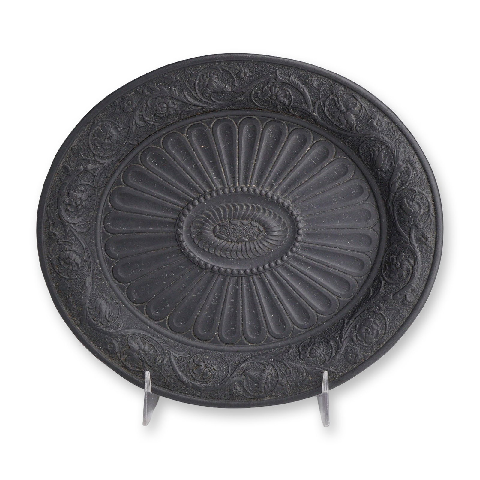 Wedgwood Black Basalt Oval Plate: The item is composed of fine-grained, unglazed black stoneware. The shallow form features an elaborately relief-molded upper surface. The central well is decorated with radiating flutes or gadrooning,
