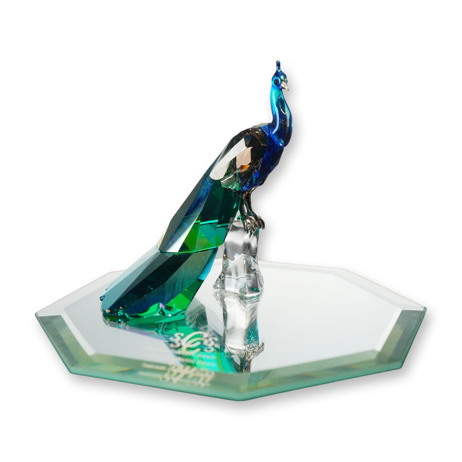 Swarovski Crystal SCS Loyalty Figurine, Peacock: The bird is depicted perched on a clear, craggy crystal base, featuring a faceted deep blue neck and body, with a faceted emerald green and iridescent yellow-to-green Aurora Borealis coated crystal ta