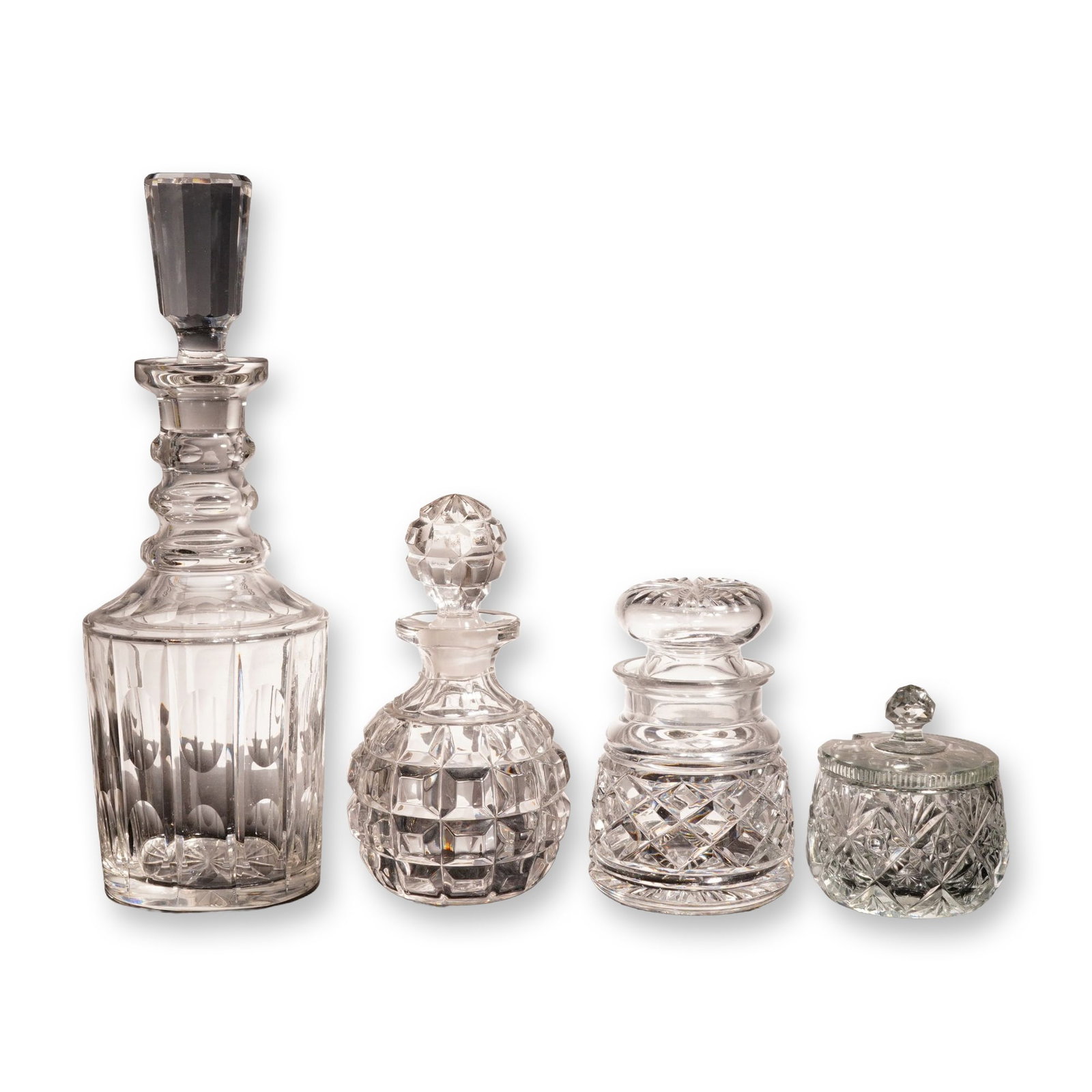 4pc Cut Crystal Decanters and Lidded Jars: The collection comprises a tall spirits decanter featuring a panel-cut neck, triple applied neck rings, and vertical flute cuts with circular thumbprints, complete with a heavy faceted hexagonal stopp