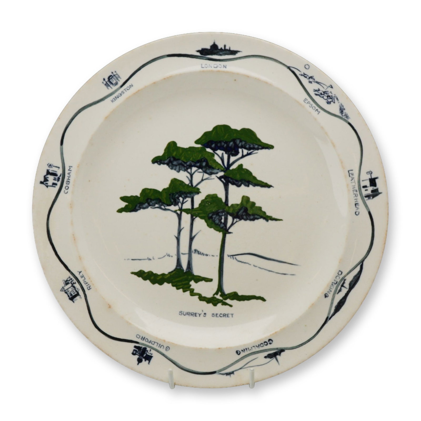 Wedgwood Etruria England Stamped Creamware Plate Featuring Surreys Secret Pattern: The central motif features three stylized green pine trees above the title Surreys Secret in black capital letters. The rim is decorated with a continuous wavy-line border depicting a travel route thr