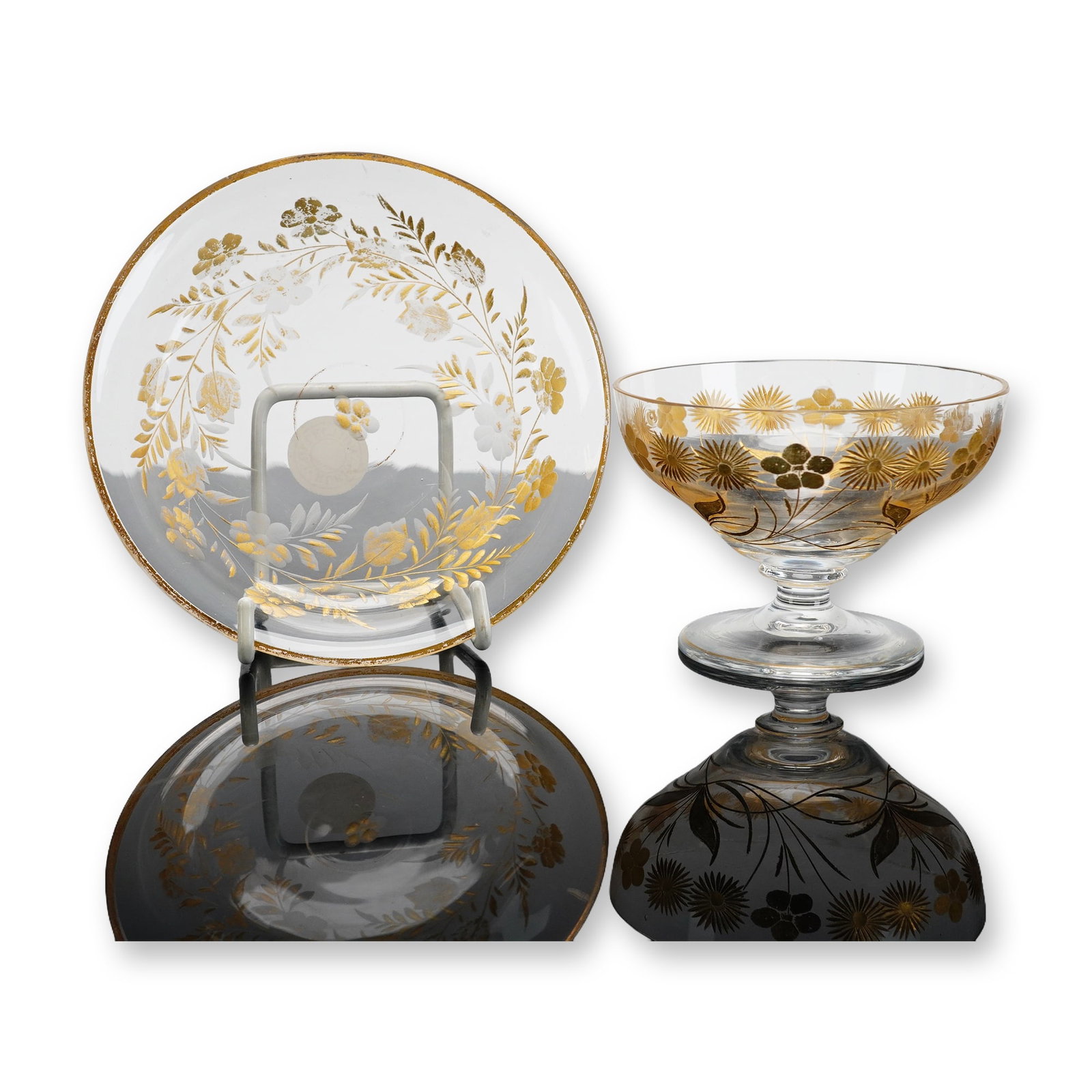 Two Clear and Gilded Glass Tableware Items: This pair of clear glass items consists of a footed dessert bowl and a matching saucer. Both pieces are decorated with ornate gilded floral and foliate motifs, including stylized sunburst flowers and 