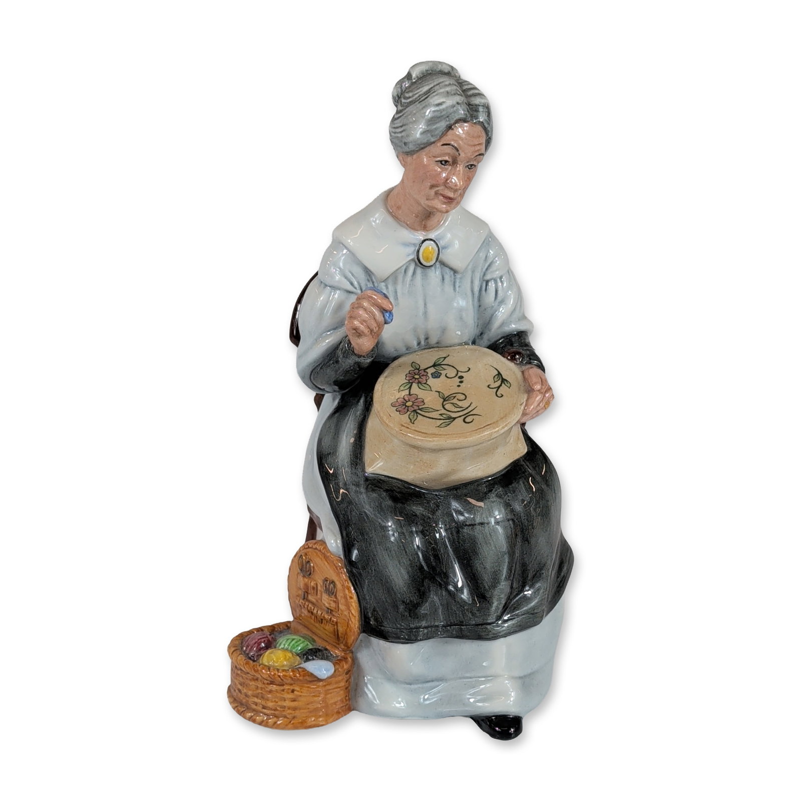 Royal Doulton Porcelain Figurine Embroidering HN2855: Royal Doulton glazed porcelain figurine titled Embroidering, model HN 2855. The figure depicts an elderly woman seated in a wood-framed armchair with light green upholstery, holding a circular embroid