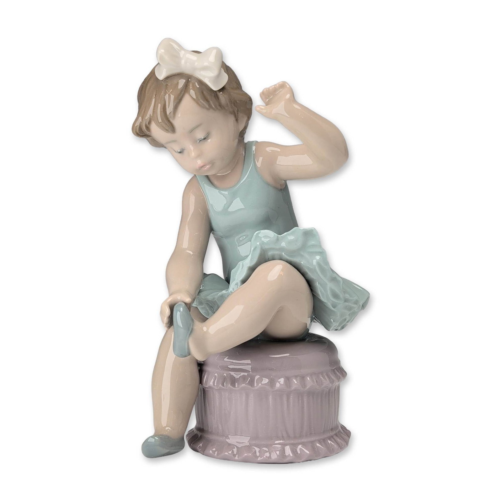 Lladro Spanish Porcelain Little Ballet Girl Figurine 5107: Lladro, Spain. Glazed porcelain figurine titled Little Ballet Girl, model number 5107. Features a seated young ballerina dressed in a pale blue leotard and ruffled tutu atop a lavender fluted pedestal