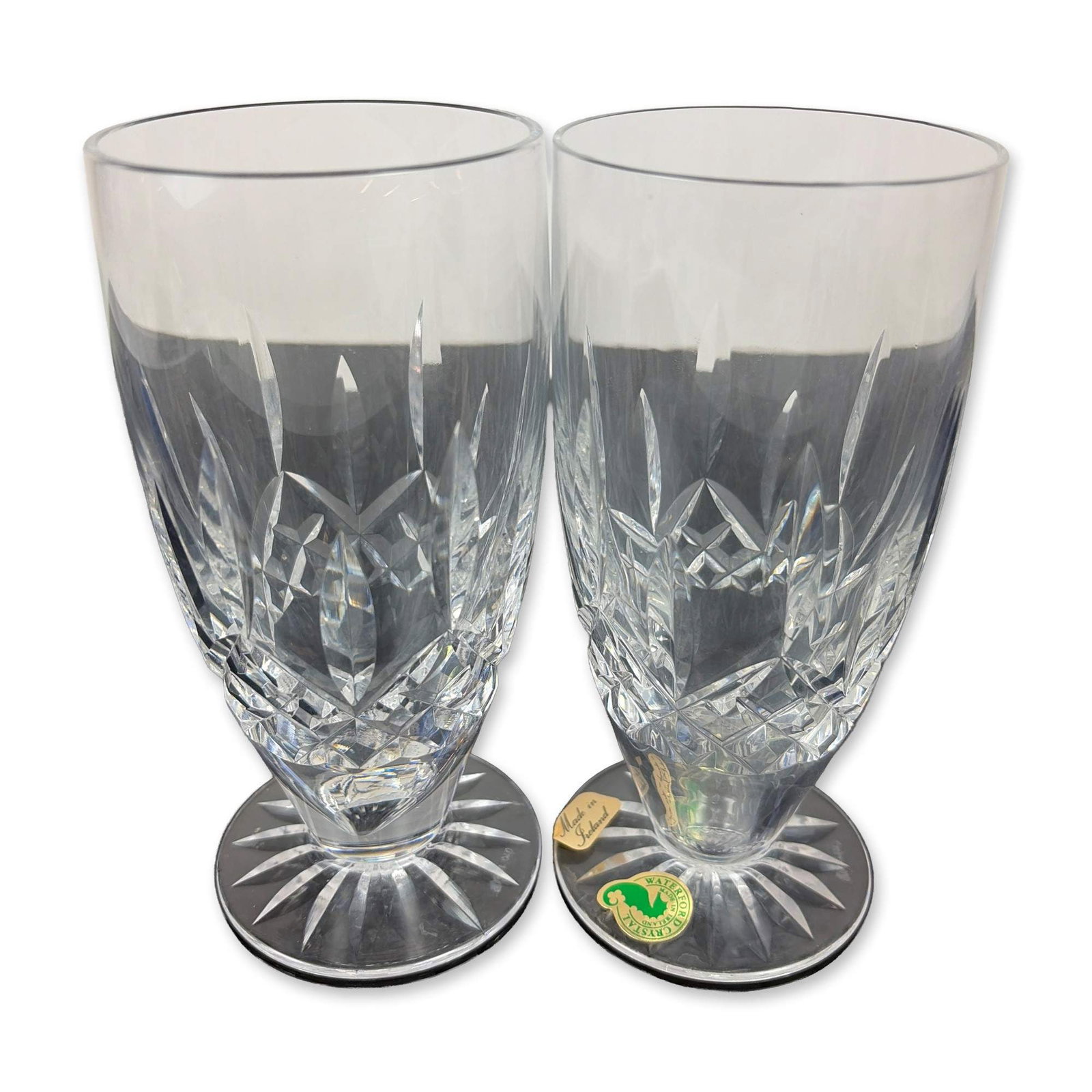 Pair of Waterford Lead Crystal Lismore Footed Iced Tea Glasses: Heavy lead crystal construction featuring a central band of diamond cuts flanked by vertical wedge cuts. The base of each glass exhibits a multi-point radial star cut. Both pieces bear the acid-etched