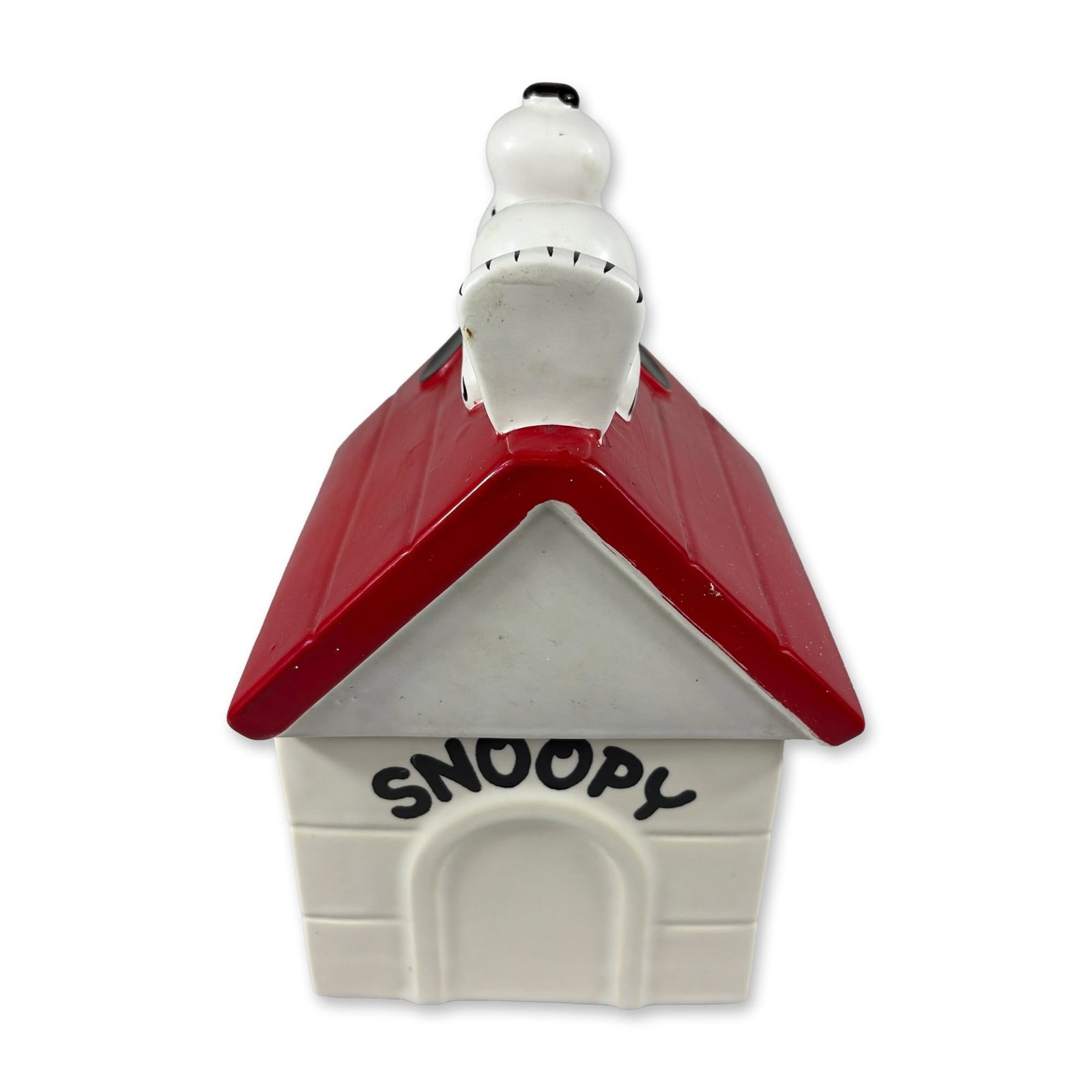 1970 Peanuts Snoopy Doghouse Ceramic Cookie Jar by United Feature Syndicate: This two-piece glazed ceramic cookie jar is modeled as Snoopy's iconic red-roofed doghouse, featuring the character lying on his back across the gabled peak of the lid. The white square base includes 