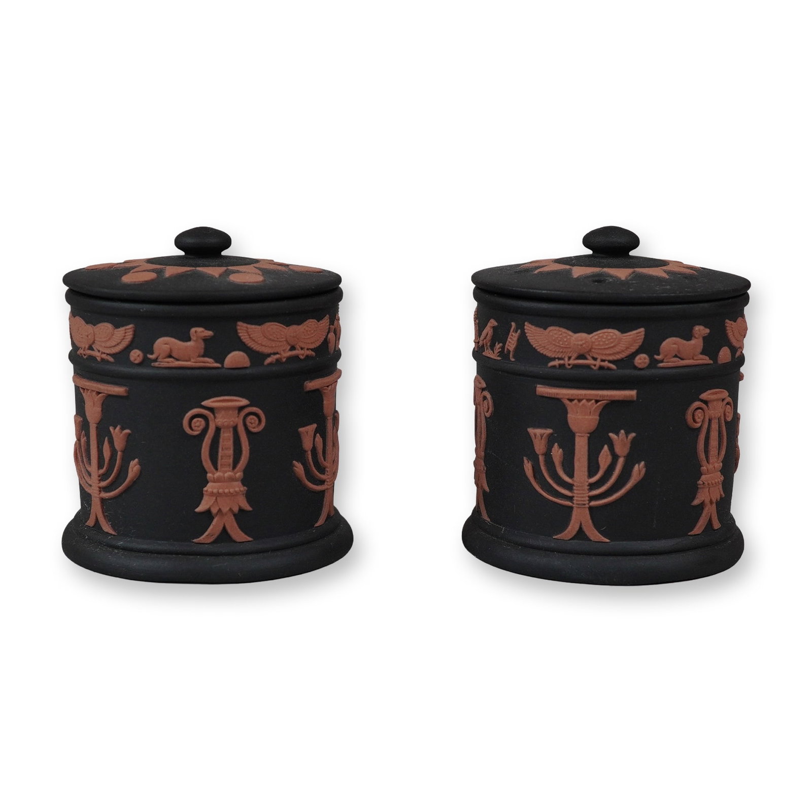 2pc Wedgwood Black Basalt and Terracotta Egyptian Revival Potpourri Jars: This lot consists of a pair of matching cylindrical jars with lids manufactured by Wedgwood in England. Each piece is crafted from matte black basalt stoneware and features terracotta-colored applied 