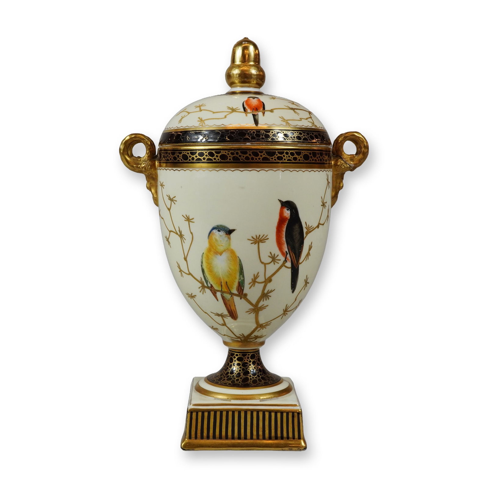 Wedgwood Bone China Gilt Paste and Enameled Bird Covered Urn: The neoclassical ovoid vessel features a domed lid topped with a gilded finial and is flanked by two gilded scroll handles. The urn rests on a waisted socle atop a square pedestal base. The central bo