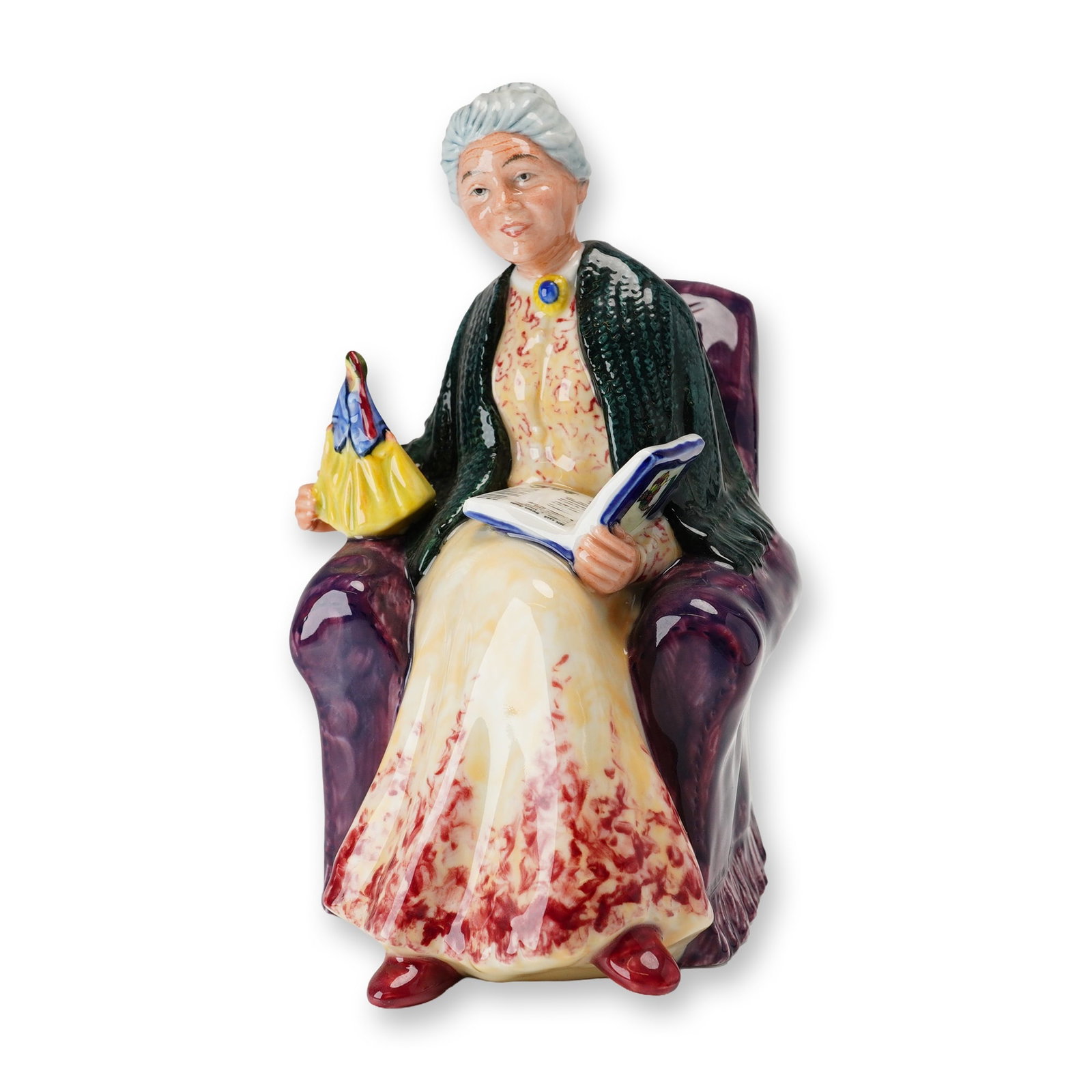 Royal Doulton Porcelain Figurine, Prized Possession HN2942: Part of the RDICC series, designed by Robert Tabbenor. Hand-painted figure depicting a seated woman in a plush purple armchair, wrapped in a green shawl. She holds a Royal Doulton figurine in one hand