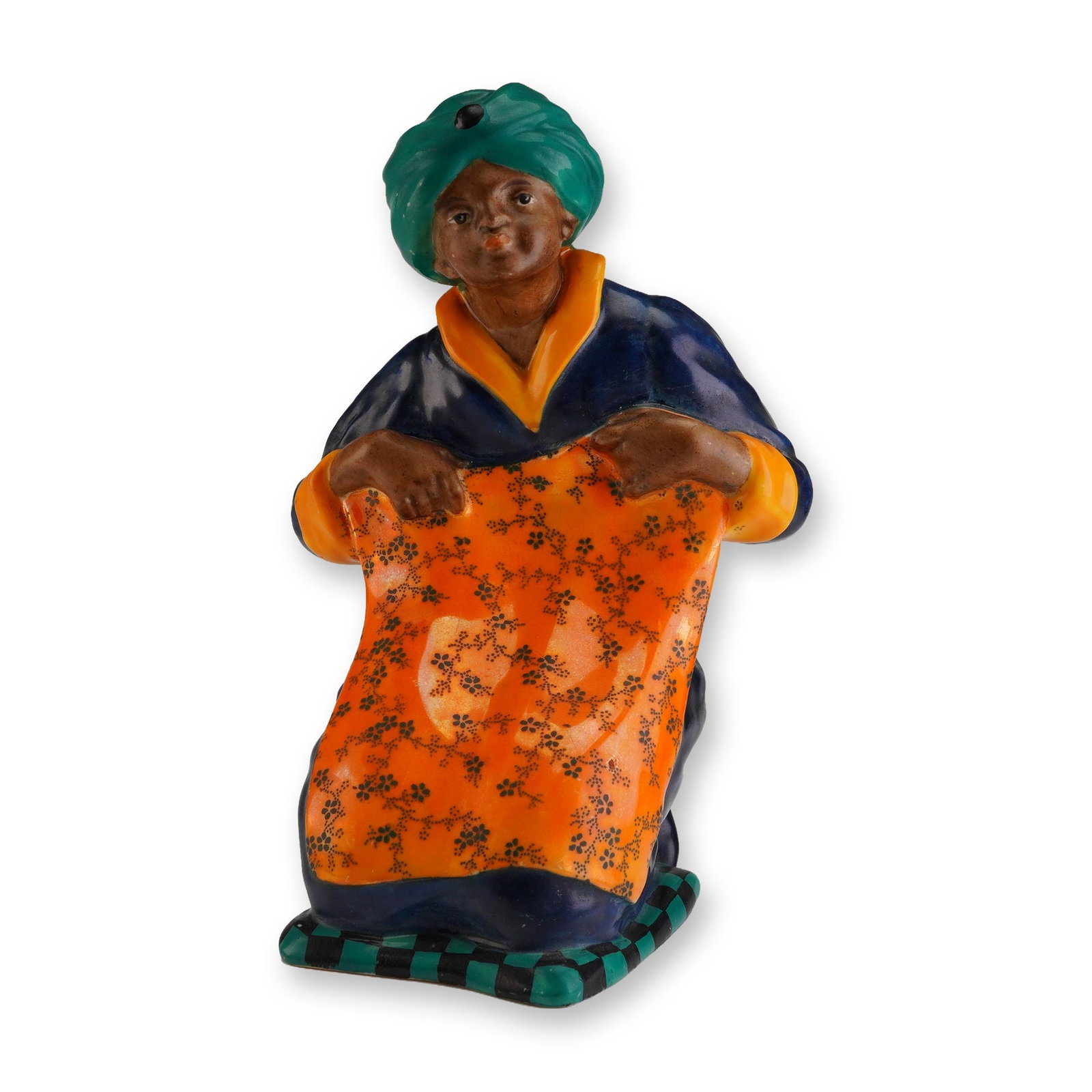 Royal Doulton Figurine, The Carpet Vendor HN76: A polychrome glazed earthenware figurine, titled The Carpet Vendor, designed by Charles J. Noke. The figure is depicted kneeling, wearing a teal turban and dark blue robes trimmed in orange, holding a