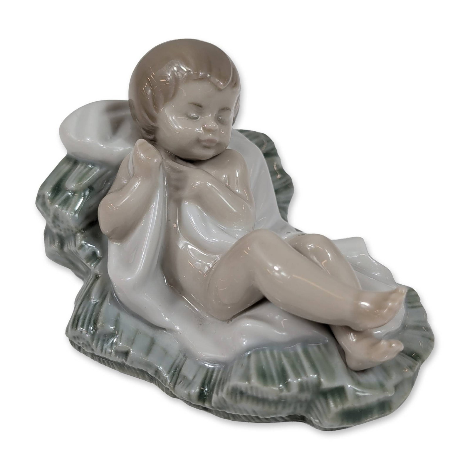 Lladro Porcelain Baby Jesus Figurine 5478: Lladro (Spain). Nativity figure of Baby Jesus in glazed porcelain, depicting a sleeping infant partially draped in a white cloth and resting on a textured green bed of straw. The base bears the blue p