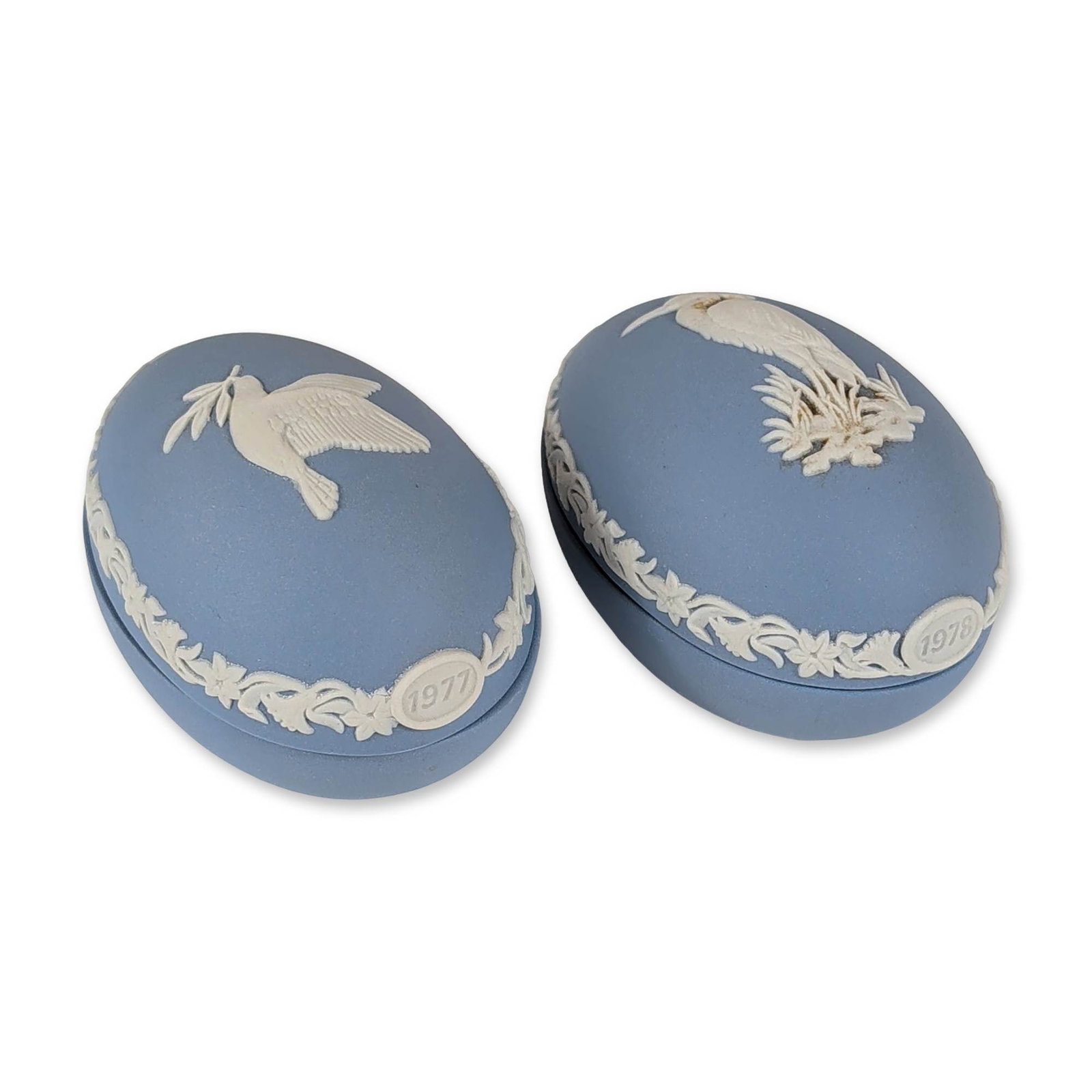 Pair Wedgwood Jasperware Easter Egg Trinket Boxes: Wedgwood, England. Pair of egg-shaped lidded trinket boxes in blue jasperware with white bas-relief application. One example is dated 1977 and depicts a dove with an olive branch, while the second is 