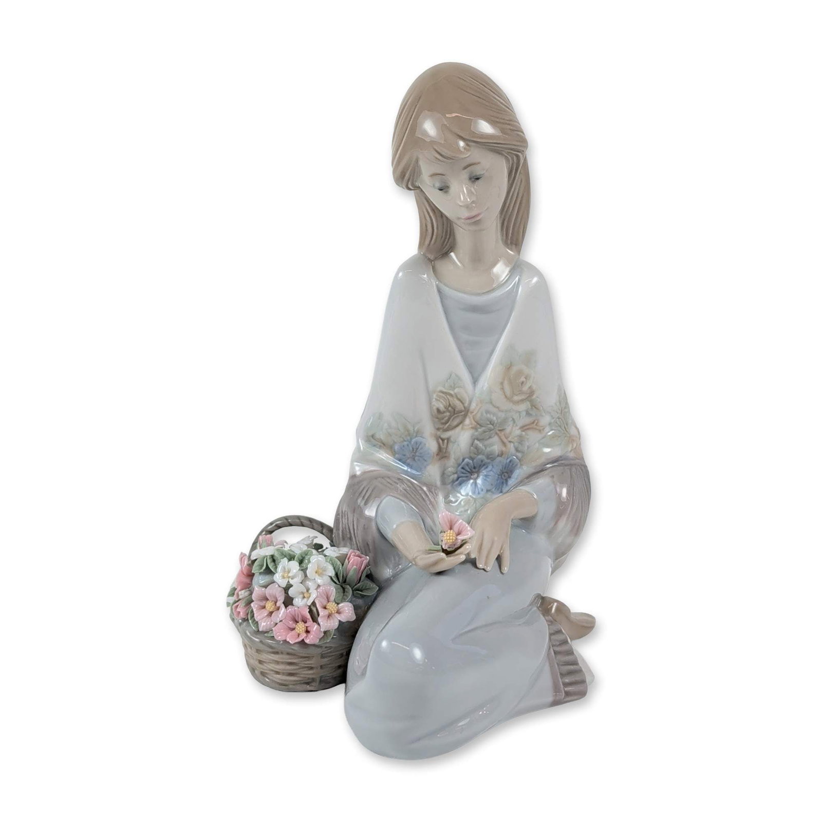 Lladro Porcelain Figurine, Flower Song 1007607: The Lladro glazed porcelain figurine titled Flower Song, model number 7607, was designed by Juan Huerta. It features a seated girl in a shawl holding a single pink flower, with a woven basket of assor