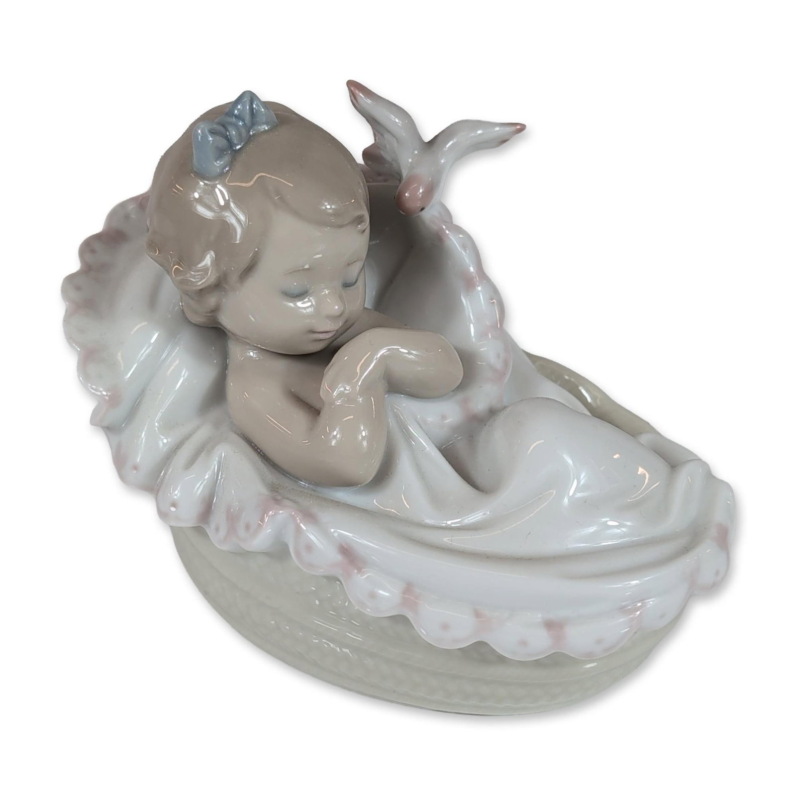 Lladro Glazed Porcelain Figurine, Comforting Dreams 01006710: Lladro (Spain). Comforting Dreams glazed porcelain figurine, model number 6710. The work depicts a sleeping infant girl in a basket with a ruffled pink-trimmed liner, featuring a blue bow in her hair 