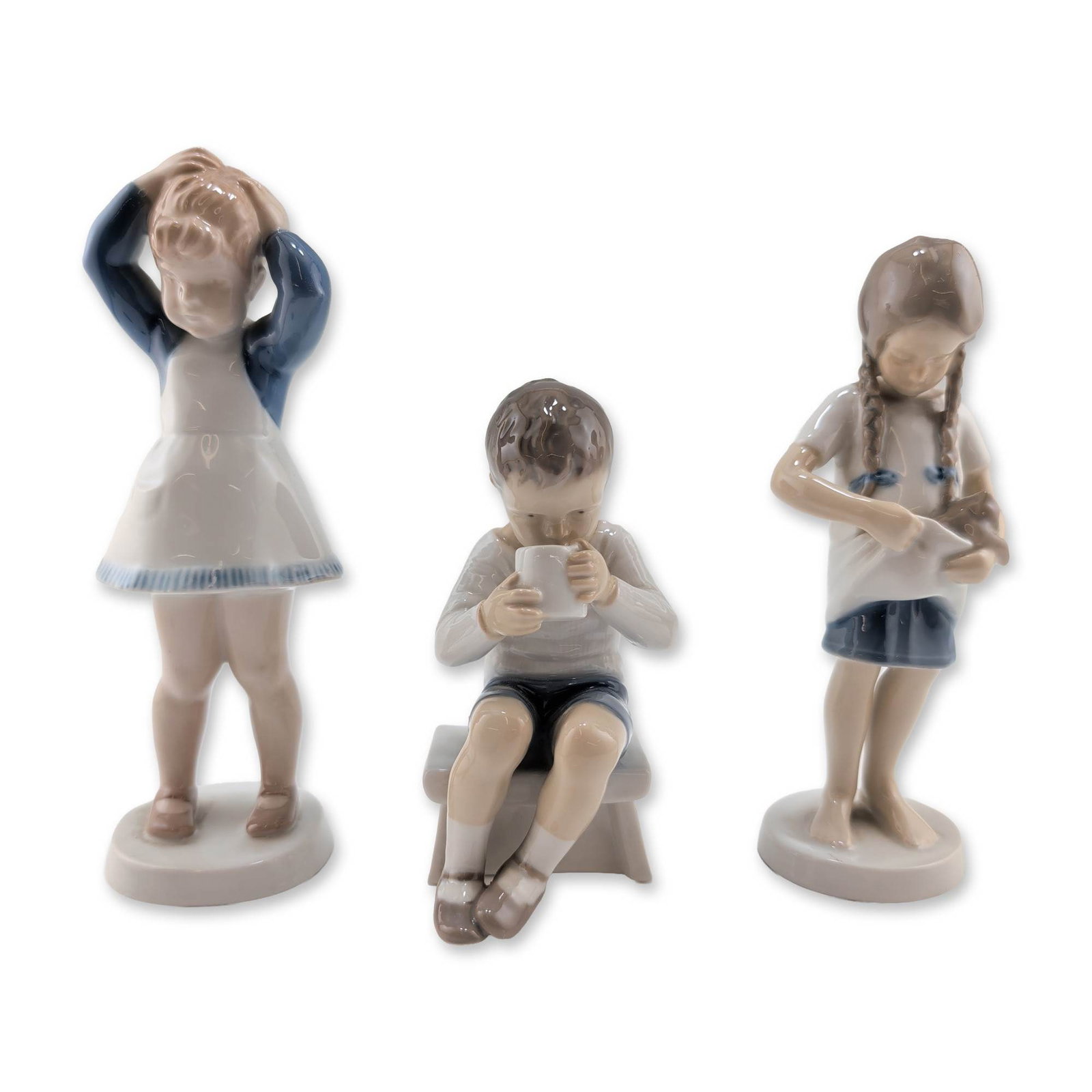 Three Bing & Grondahl Danish Porcelain Child Figurines: Bing & Grondahl, Denmark. Trio of glazed porcelain figurines depicting child subjects. The collection includes model 1779 featuring a girl with braids holding a kitten, model 1713 depicting a boy seat