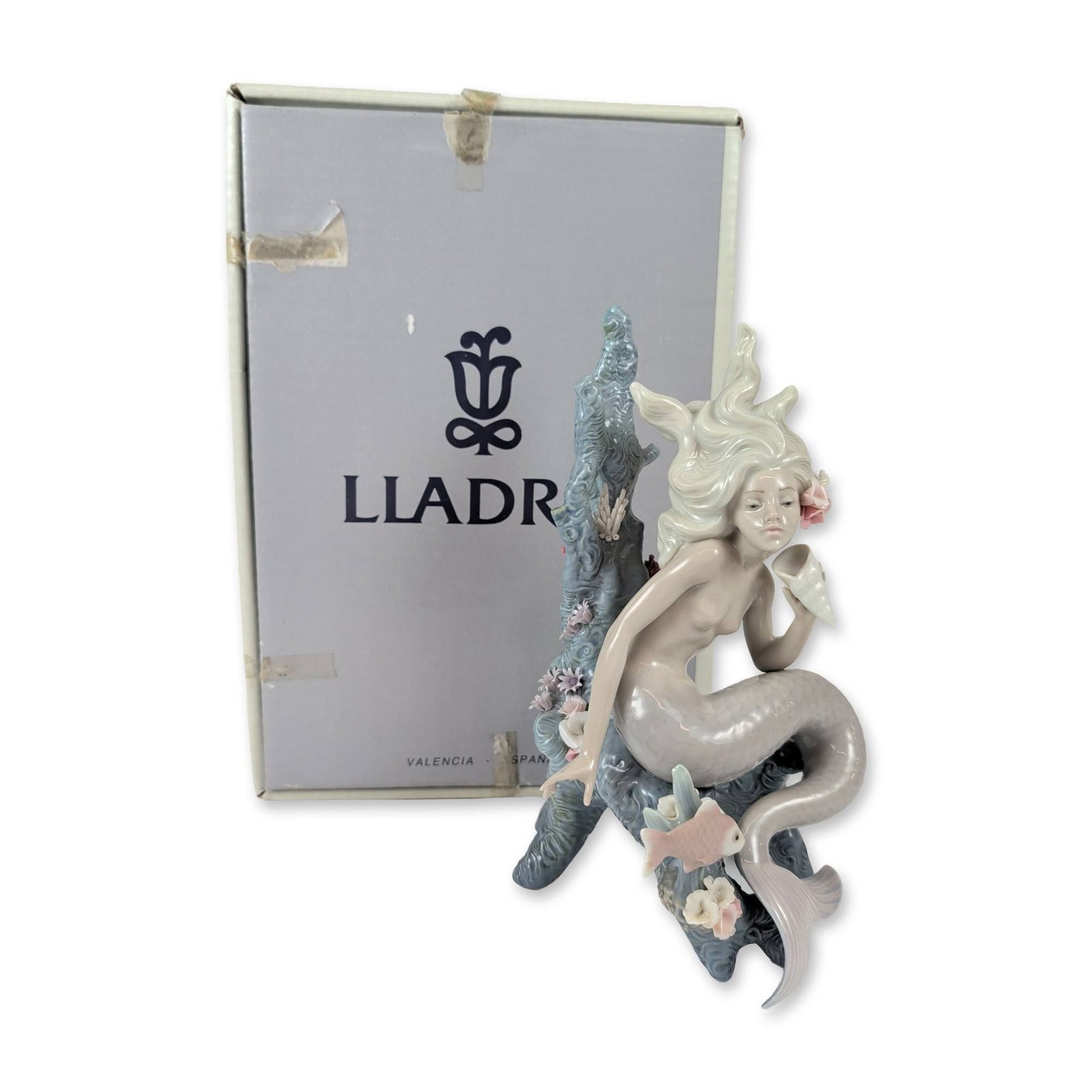 Lladro Ocean Beauty Porcelain Mermaid Figurine: Lladro (Spain). Ocean Beauty figurine, model 5785, in glazed porcelain. The figure depicts a mermaid seated on a blue-grey coral base, holding a white conch shell to her ear. Details include flowing p