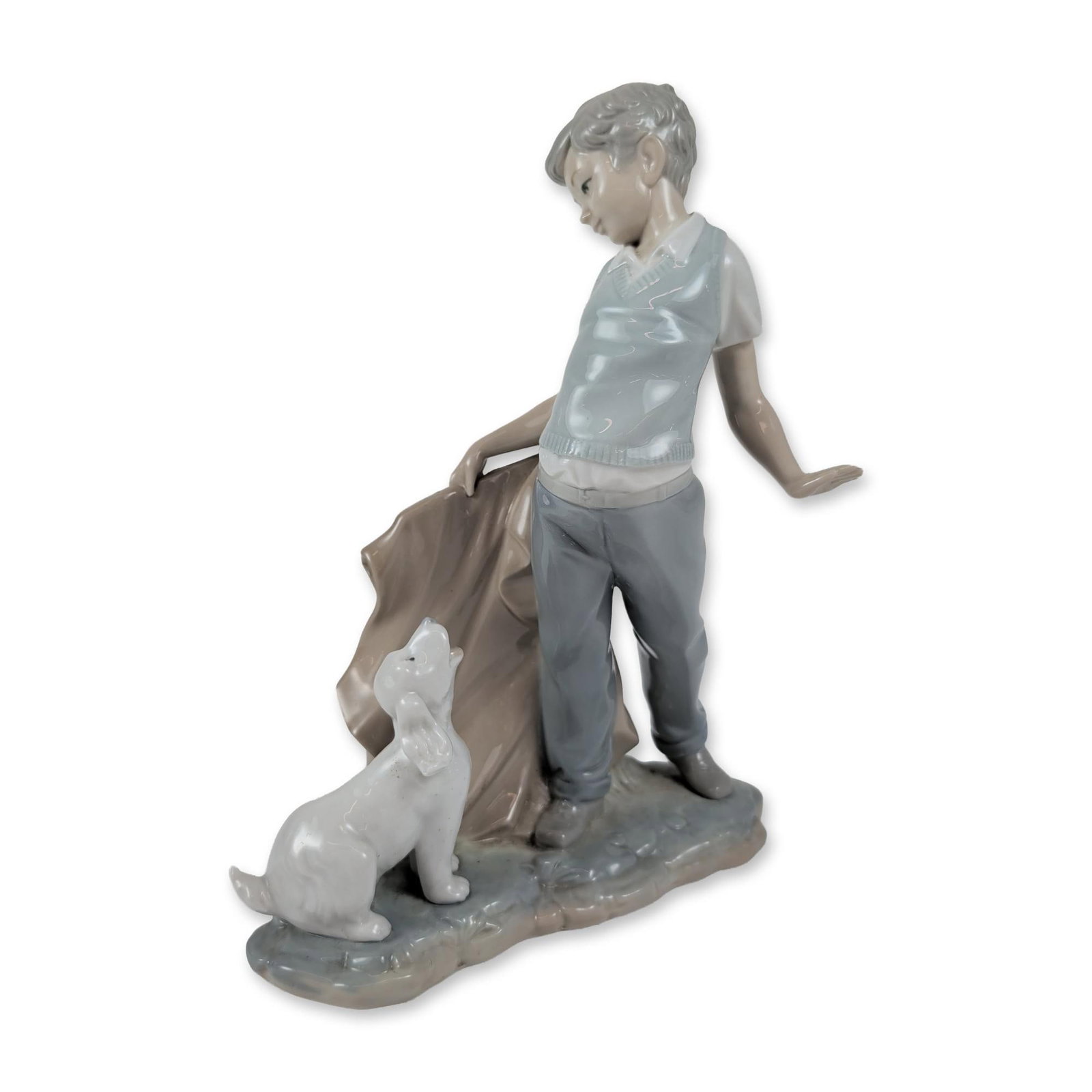 NAO by Lladro Fighting the Dog Porcelain Figurine: The sculpture depicts a boy in a light blue sweater vest and gray trousers holding a piece of draped fabric, with one hand raised toward a small white dog at his feet. The underside is marked with the