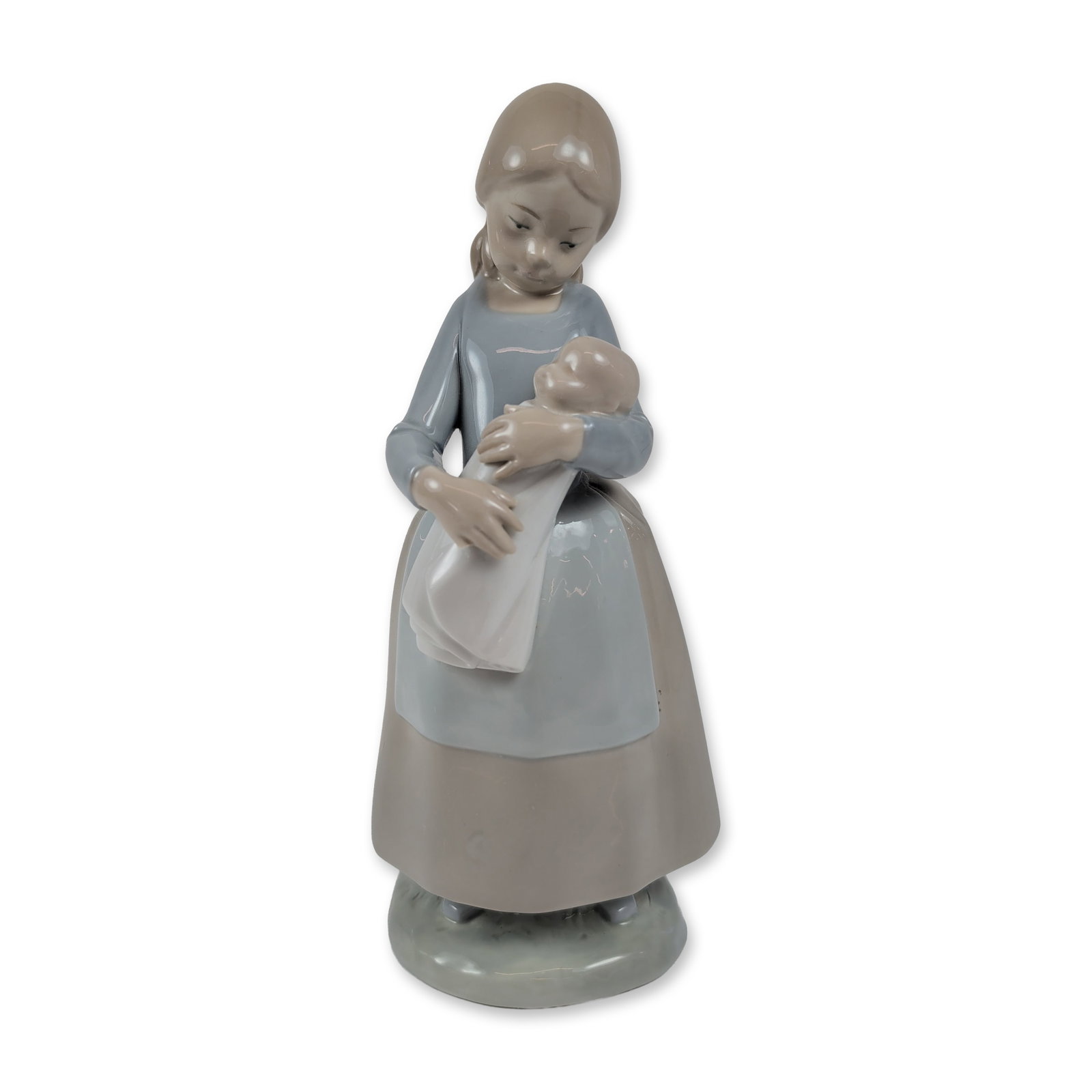 NAO by Lladro Porcelain Candid Figurine: NAO by Lladro, Spain. Glazed porcelain figurine depicting a standing girl cradling a swaddled infant or doll. The figure is dressed in a light blue long-sleeved blouse and a gray-tan skirt with a pale