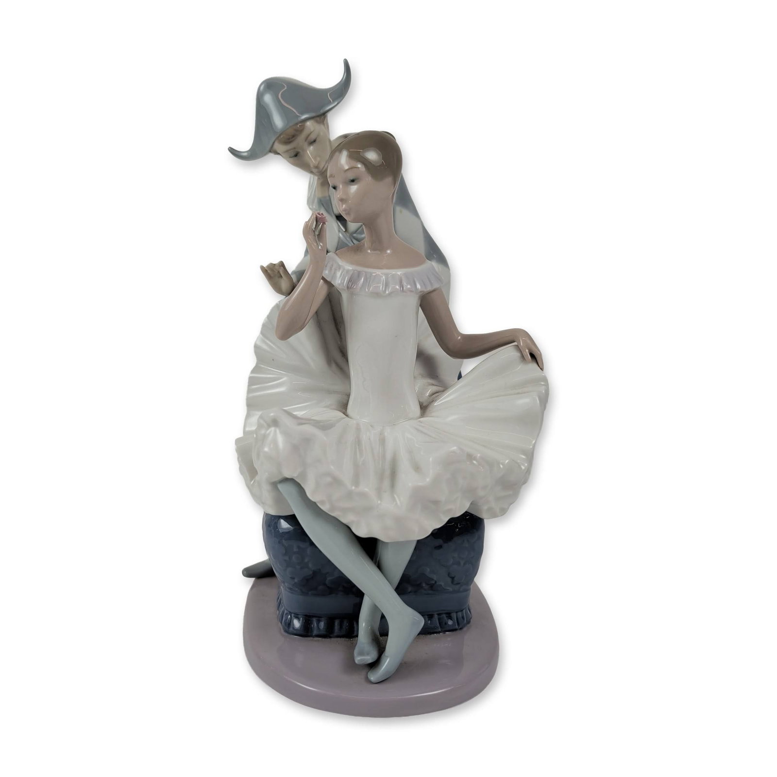 NAO by Lladro Porcelain Figurine, Harlequin and Ballerina: This piece depicts a seated ballerina in a white tutu holding a single pink flower, with a harlequin figure in a diamond-patterned bodysuit leaning behind her. They rest on a blue patterned ottoman ba