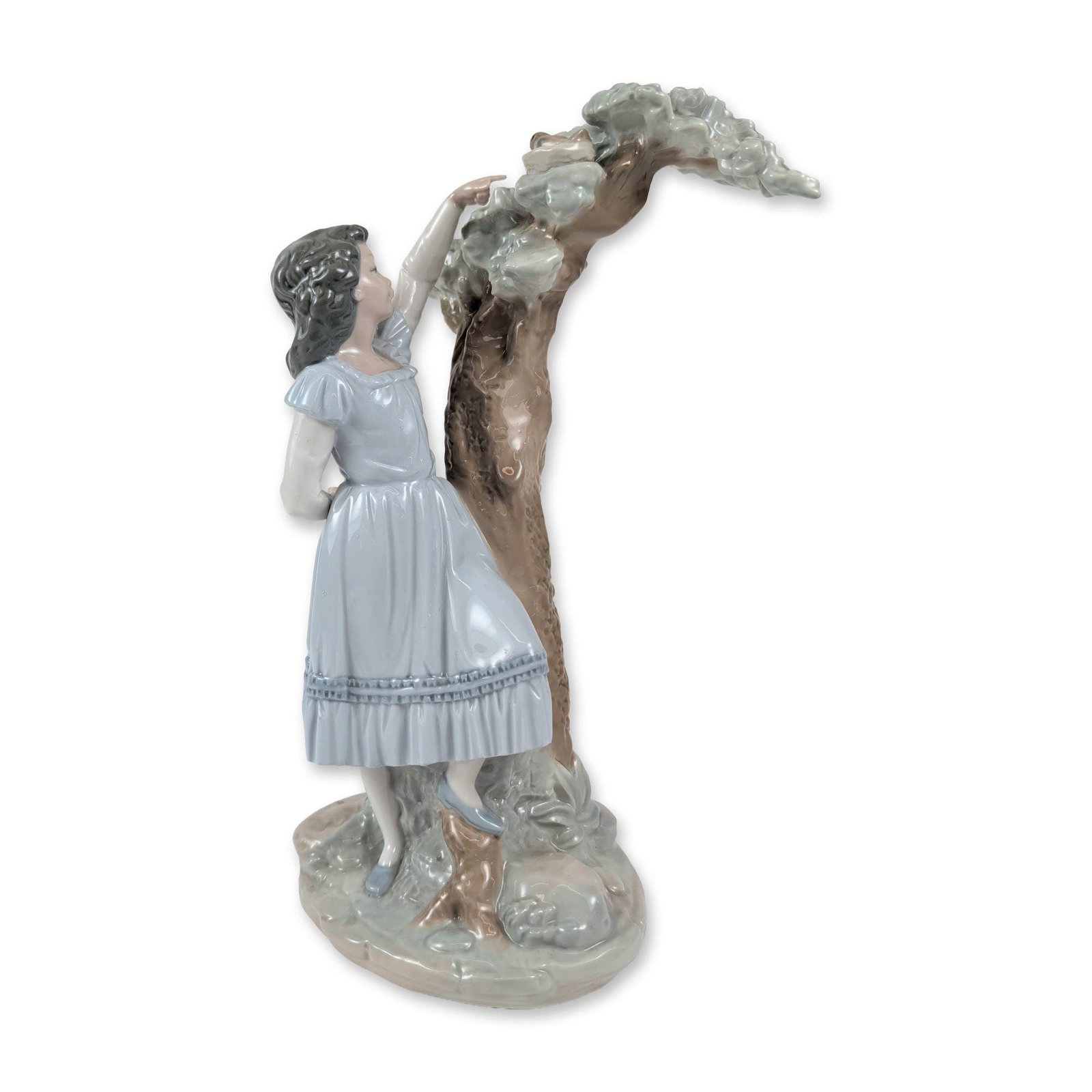 Nao by Lladro Nest Discovery Porcelain Figurine: The piece features a young girl in a pale blue short-sleeved dress with a ruffled hem, posed leaning against a gnarled tree trunk while reaching toward the upper branches. The figurine is finished in 