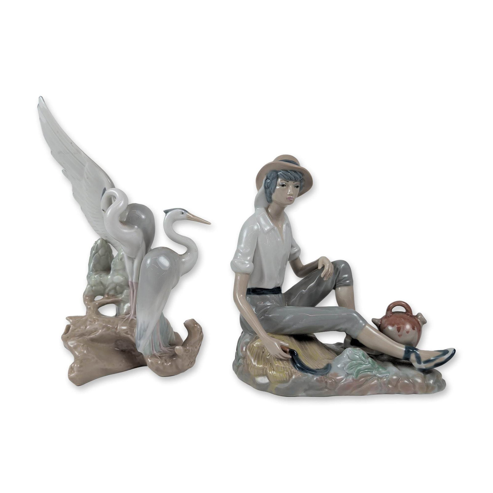 Two Spanish Porcelain Figurines, Nao by Lladro and Casades: The Casades piece portrays a young boy in rustic attire, lounging against a sheaf of wheat with a scythe in hand and a brown earthenware jug nearby. The Nao figurine showcases a graceful pair of heron