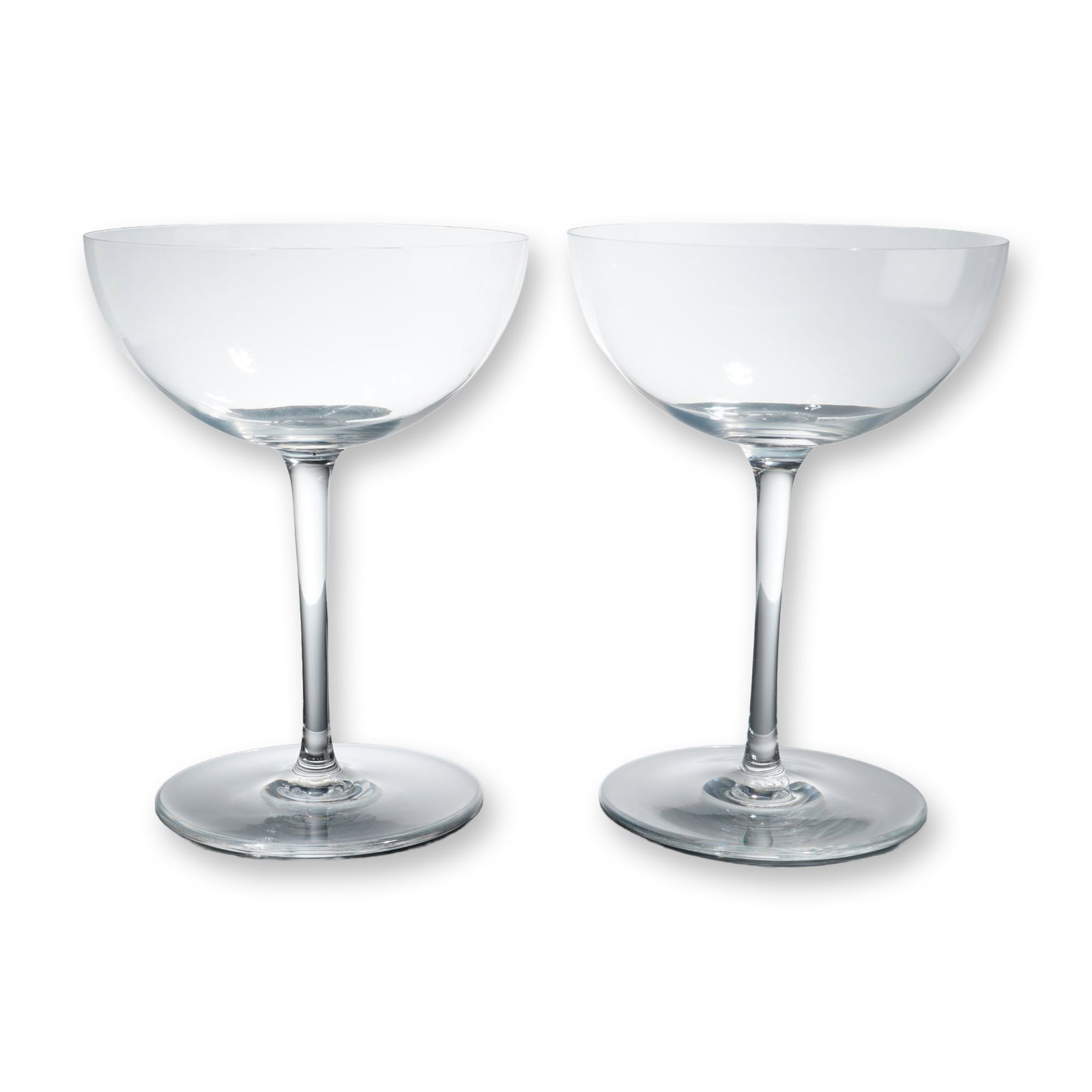 Pair of Baccarat Crystal Tall Champagne Coupes: Baccarat, France. Pair of tall champagne coupes in clear crystal, featuring wide, shallow, unadorned bowls with fine rims supported by slender cylindrical stems on flat circular bases. Each glass bear