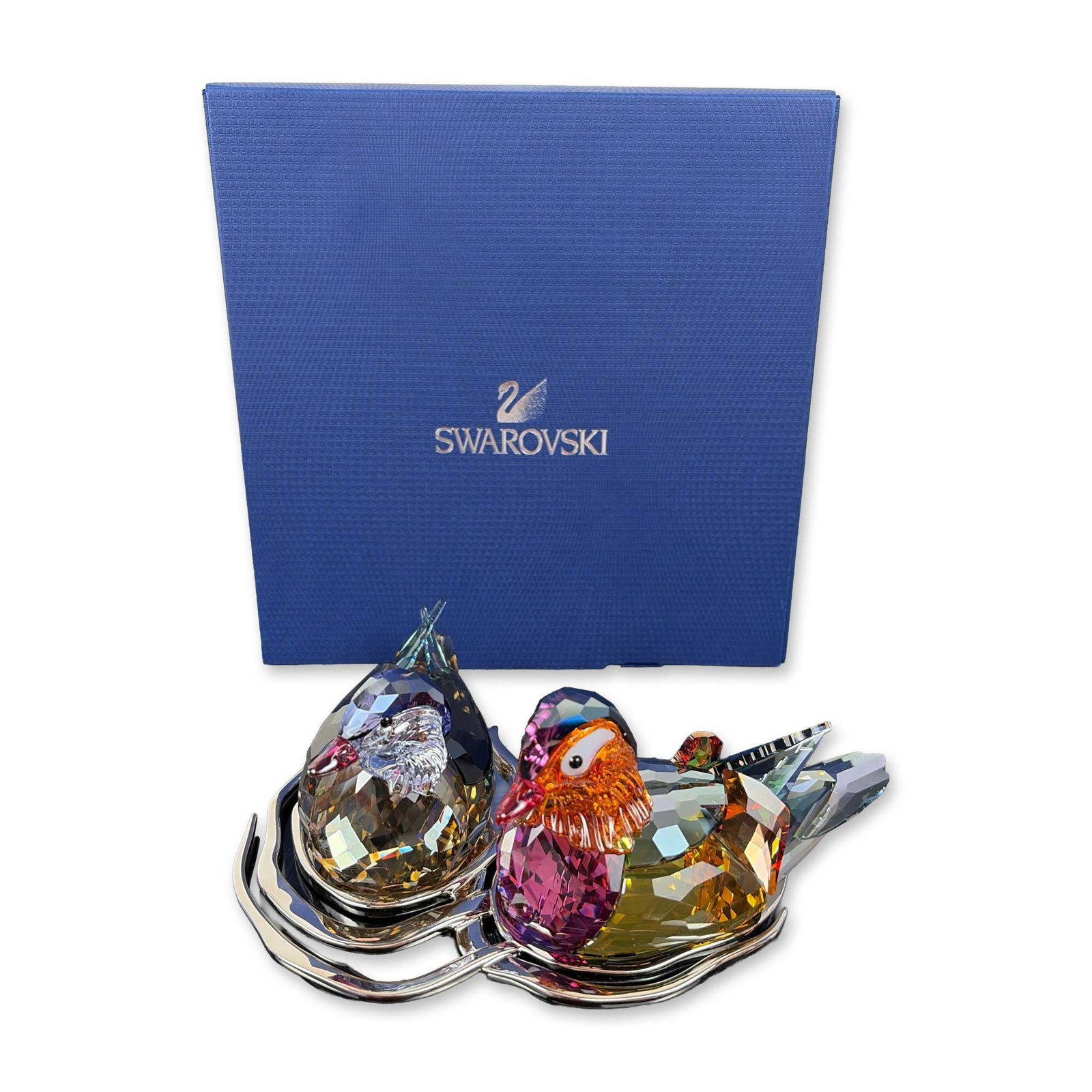 Swarovski Crystal Mandarin Ducks Figurine with Base: A pair of Mandarin ducks crafted from multi-colored faceted lead crystal, showcasing shades of amethyst, topaz, and orange. They rest on a silver-tone metal base with a smooth, wave-like design. The u