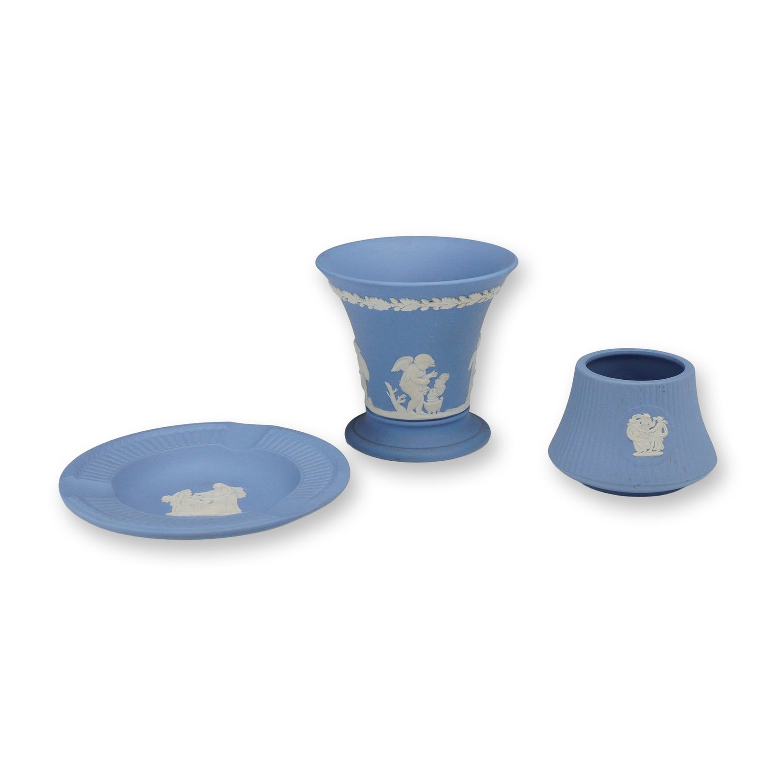 Three-Piece Wedgwood Pale Blue Jasperware Cigarette Lighter Holder, Ashtray and Vase: The first item is a cigarette lighter holder with a flared rim. It is decorated with a laurel leaf border and a central relief depicting putti at play. The second item is a small circular ashtray