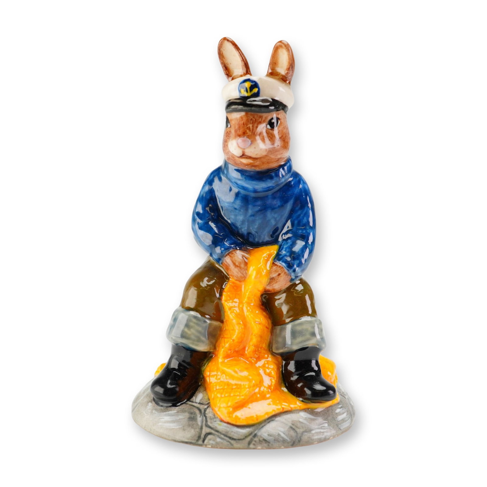 Royal Doulton Prototype Bunnykins Figurine, Fisherman: A rare Royal Doulton Bunnykins prototype figurine depicting a fisherman rabbit seated on a black bollard while mending a yellow fishing net. The figure is dressed in a dark blue sweater, white cap, kh