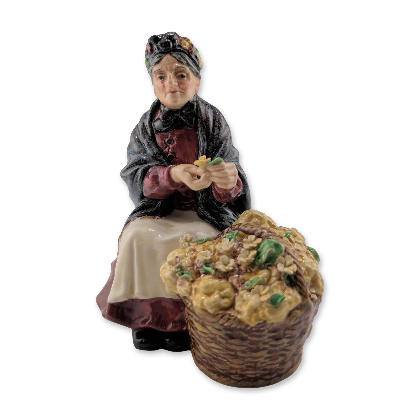 Royal Doulton Porcelain Figurine, Primroses HN1617: Hand-painted glazed porcelain depicting an elderly flower seller seated beside a woven basket filled with yellow primroses. The figure is modeled in a maroon bodice, black shawl, and cream-colored apr