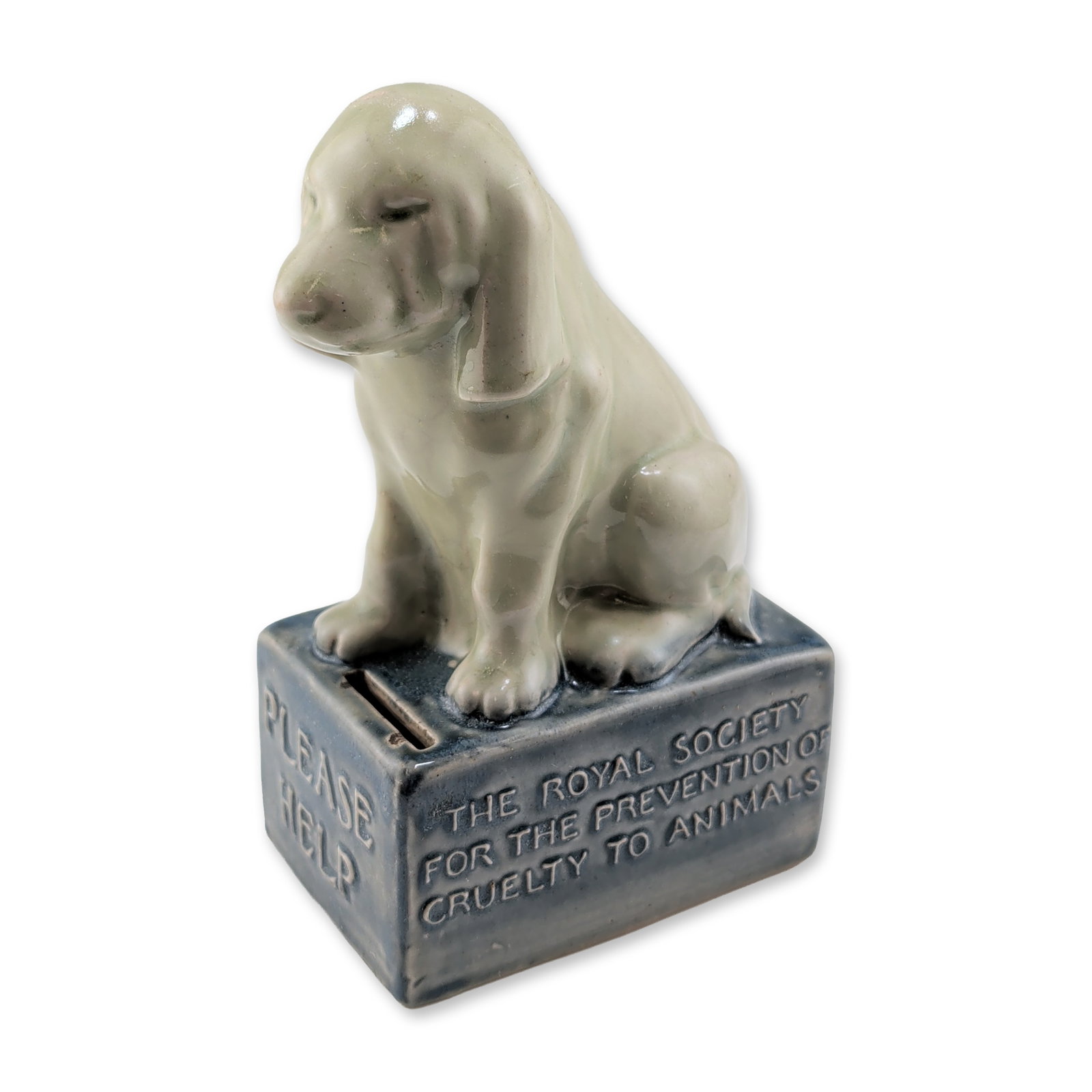 Doulton Lambeth Stoneware RSPCA Dog Collection Box: Still bank collection box in glazed stoneware, featuring a seated dog figure. The figurine is finished in a pale green glaze atop a rectangular base with a mottled dark blue-gray glaze. The front of t