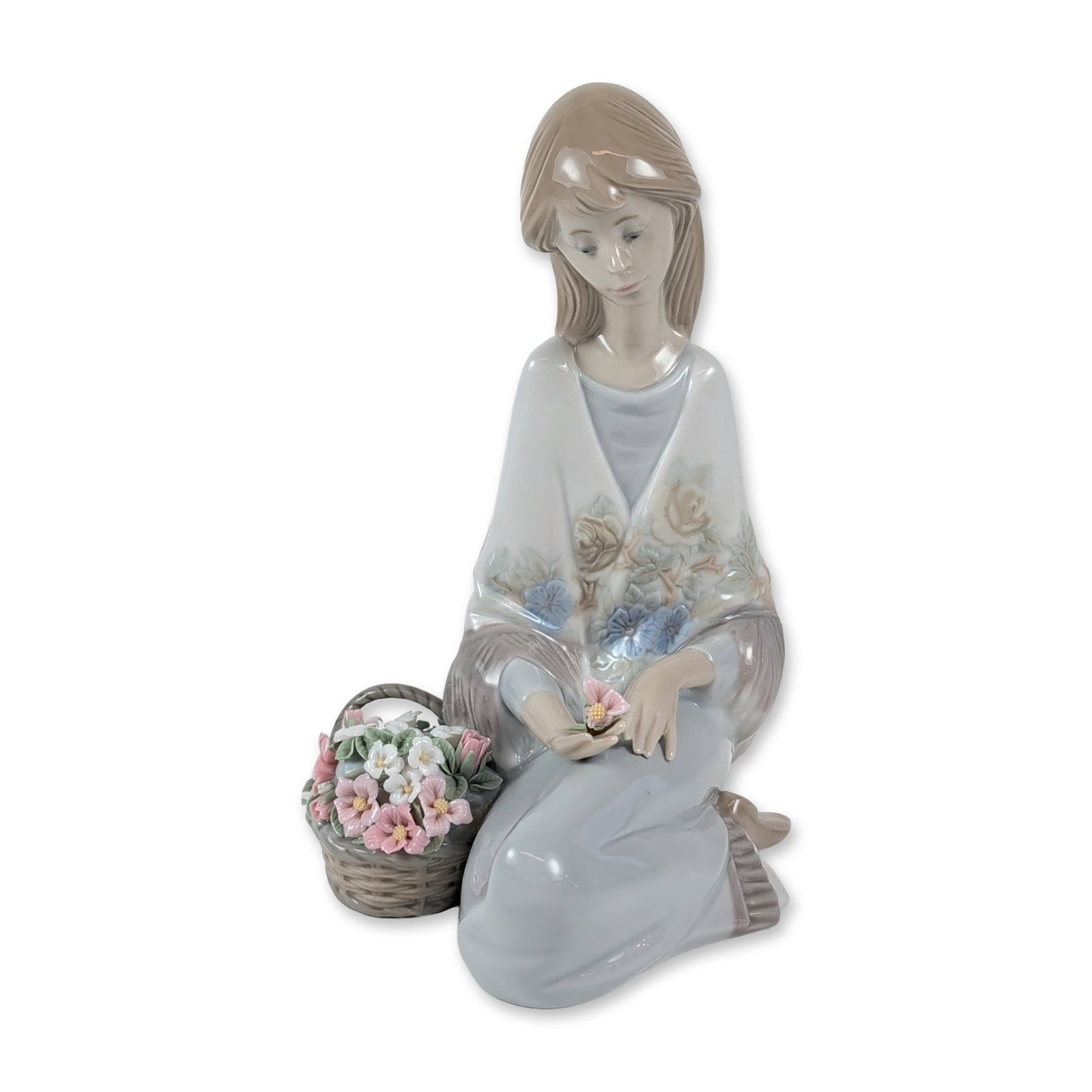 Lladro Porcelain Figurine, Flower Song 1007607: The Lladro glazed porcelain figurine titled Flower Song, model number 7607, was designed by Juan Huerta. It features a seated girl in a shawl holding a single pink flower, with a woven basket of assor