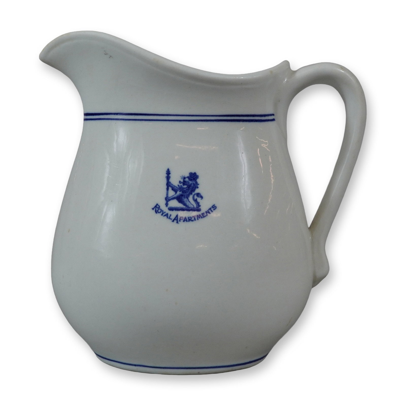 John Wanamaker Royal Apartments White China Milk Pitcher with Blue Crest: This white ceramic milk pitcher or creamer features commercial-style decoration in blue. The front is decorated with a blue transfer-printed crest depicting a lion rampant holding a standard or staff