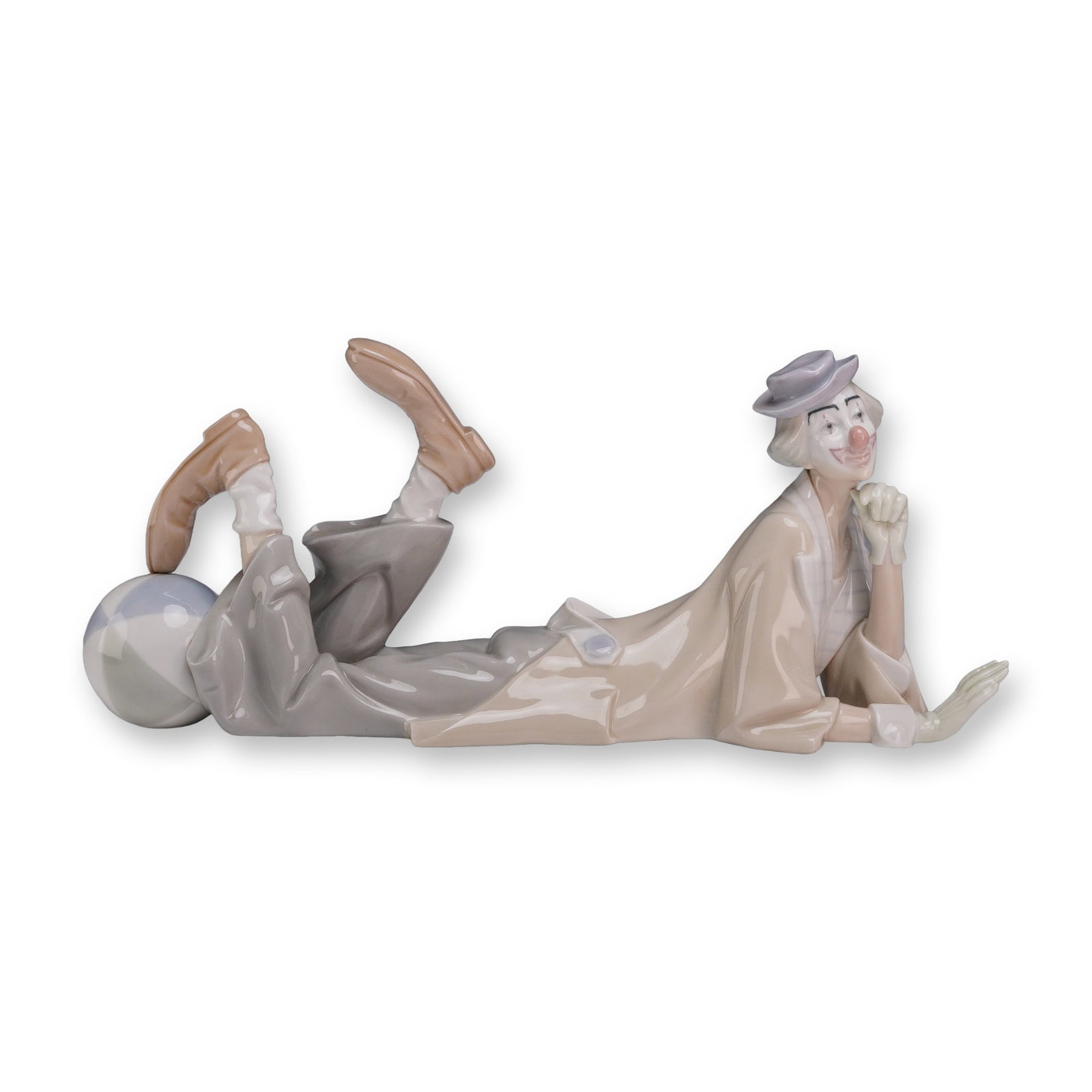 Lladro Porcelain Figurine Clown 01004618: The piece depicts a clown in a pensive reclining pose, resting on his stomach with his chin propped in his right hand. His legs are kicked upward behind him, with his left foot balanced on a large, pa