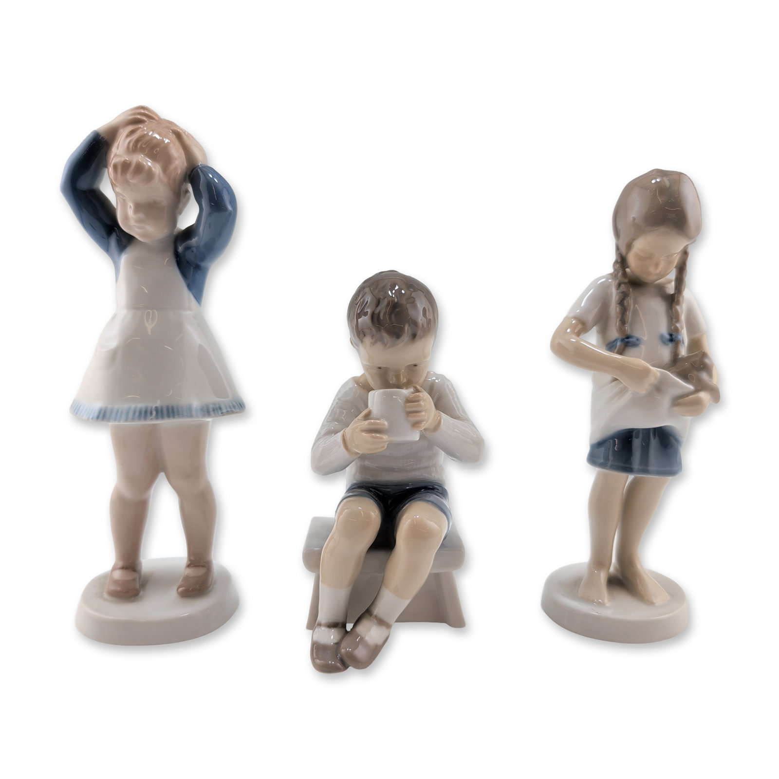 Three Bing & Grondahl Danish Porcelain Child Figurines: Bing & Grondahl, Denmark. Trio of glazed porcelain figurines depicting child subjects. The collection includes model 1779 featuring a girl with braids holding a kitten, model 1713 depicting a boy seat