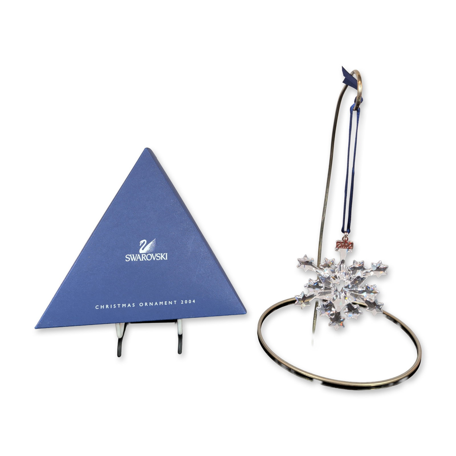 Swarovski 2004 Annual Edition Crystal Snowflake Ornament: Swarovski. 2004 Annual Edition Christmas ornament in clear faceted lead crystal, featuring a multi-pointed snowflake form. The ornament is suspended from a blue satin ribbon with an attached rectangul