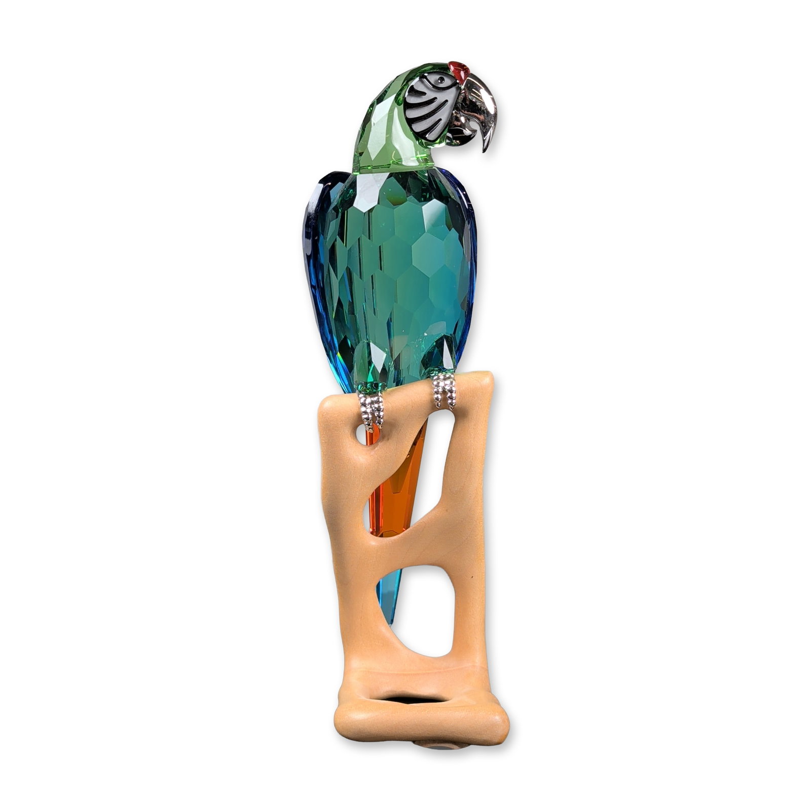 Swarovski Crystal Paradise Birds Figurine, Macaw Chrome Green: Faceted crystal parrot featuring a teal and green body, orange and blue tail, and a silver-tone metal beak with a black and white striped face panel. Perched on a matte-finished light-brown branch bas
