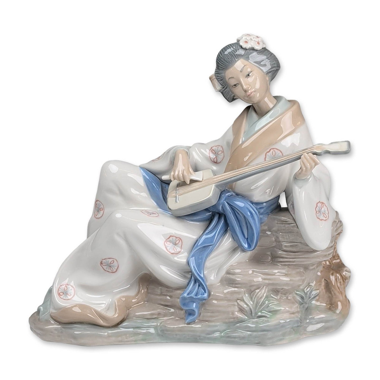 Nao by Lladro Porcelain Figurine, Geisha With Shamisen: Glazed porcelain figurine titled Geisha with Shamisen. The figure depicts a seated woman in a white kimono with pink and blue floral patterns and a large blue sash, playing a traditional instrument on