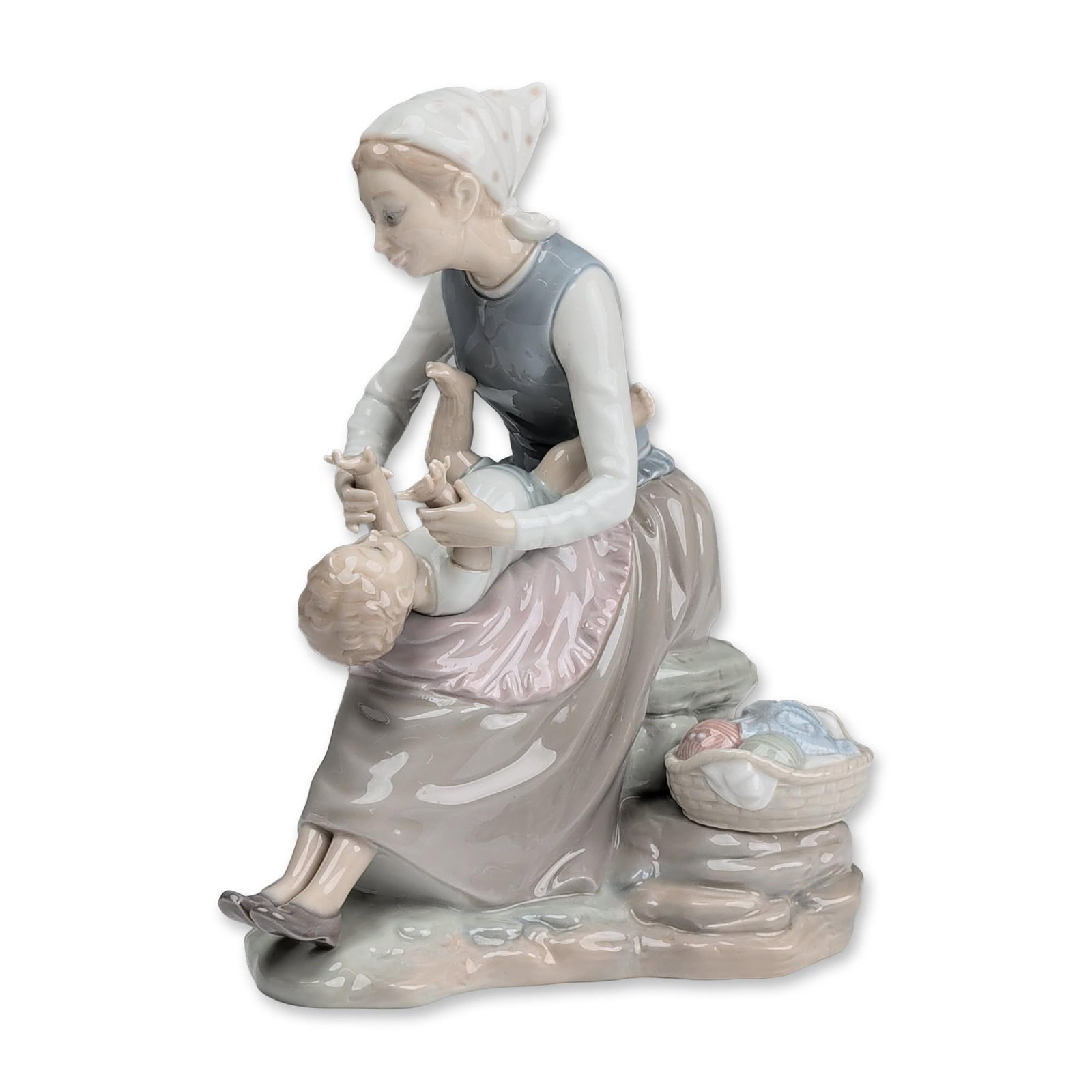 Nao Glazed Porcelain Mother and Child Figurine: Porcelain figurine in a high-gloss glaze, featuring a seated woman in rustic attire with a headscarf holding a child aloft. The figures are positioned on a rocky base alongside a basket containing lau