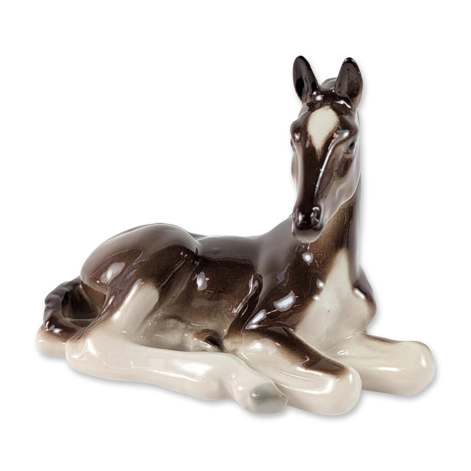 Lomonosov Russian Glazed Porcelain Foal Figurine: Lomonosov Porcelain Factory figurine of a reclining foal in glazed porcelain. The figure features a dark brown body with a white facial blaze and white markings on the legs and underside. Finished wit