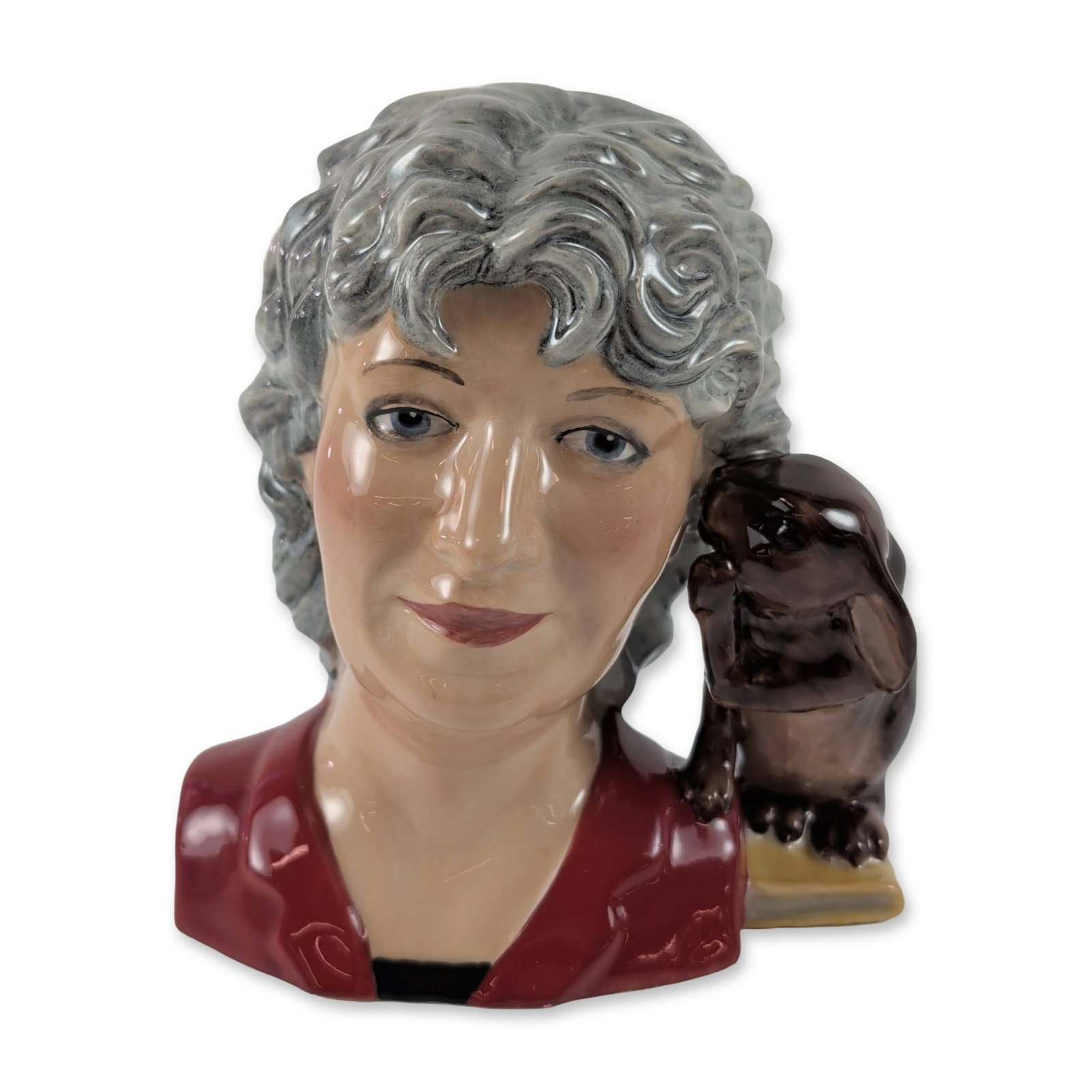 Peggy Davies Studios Louise Irvine Character Jug: Peggy Davies Studios character jug in glazed ceramic, depicting ceramic historian Louise Irvine. The figure is modeled with grey hair and a red garment, featuring a dark brown rabbit on a book resting