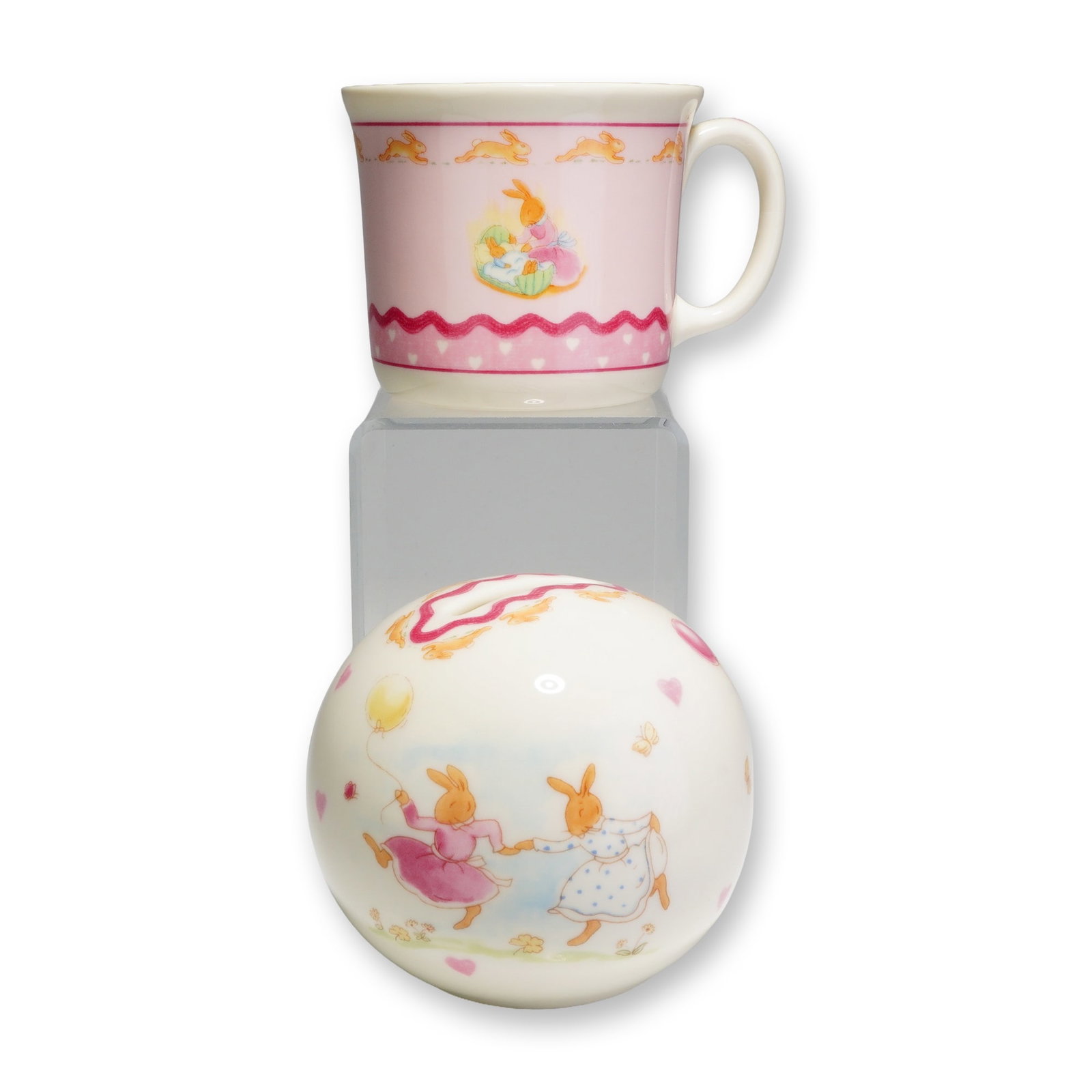2pc Royal Doulton Bunnykins Sweet Hearts Mug and Money Bank: The handled mug and spherical money box are crafted from bone china with a cream-colored ground, while the mug features a prominent pink area as its main background. Both pieces showcase colorful illu
