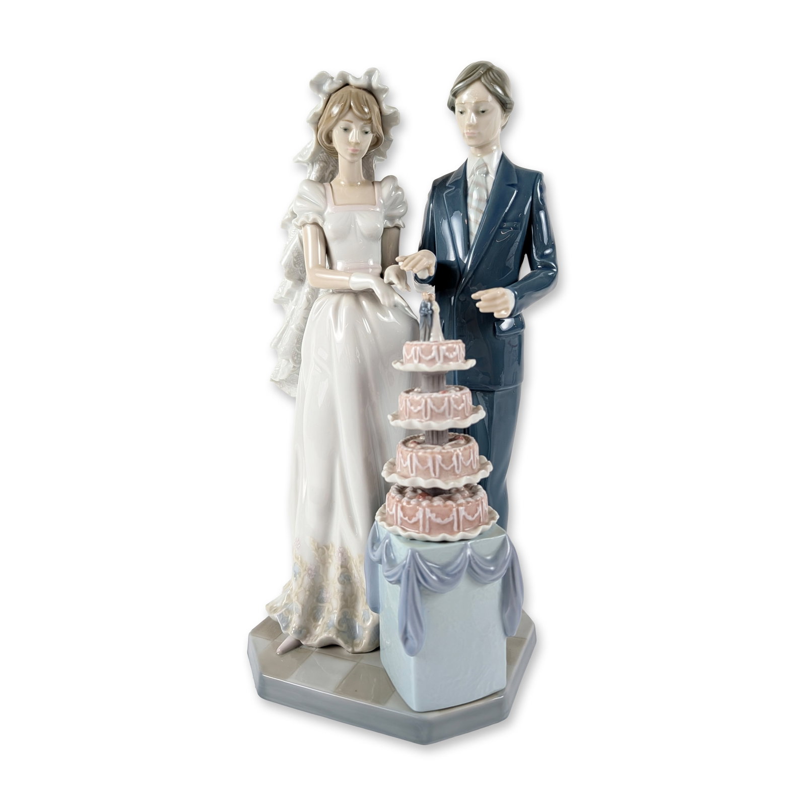 Lladro Porcelain Figurine, Wedding Cake 1005587: The figure depicts a bride and groom standing behind a four-tiered wedding cake mounted on a draped pedestal. The bride is dressed in a white gown with a textured lace-style veil and floral hem detail