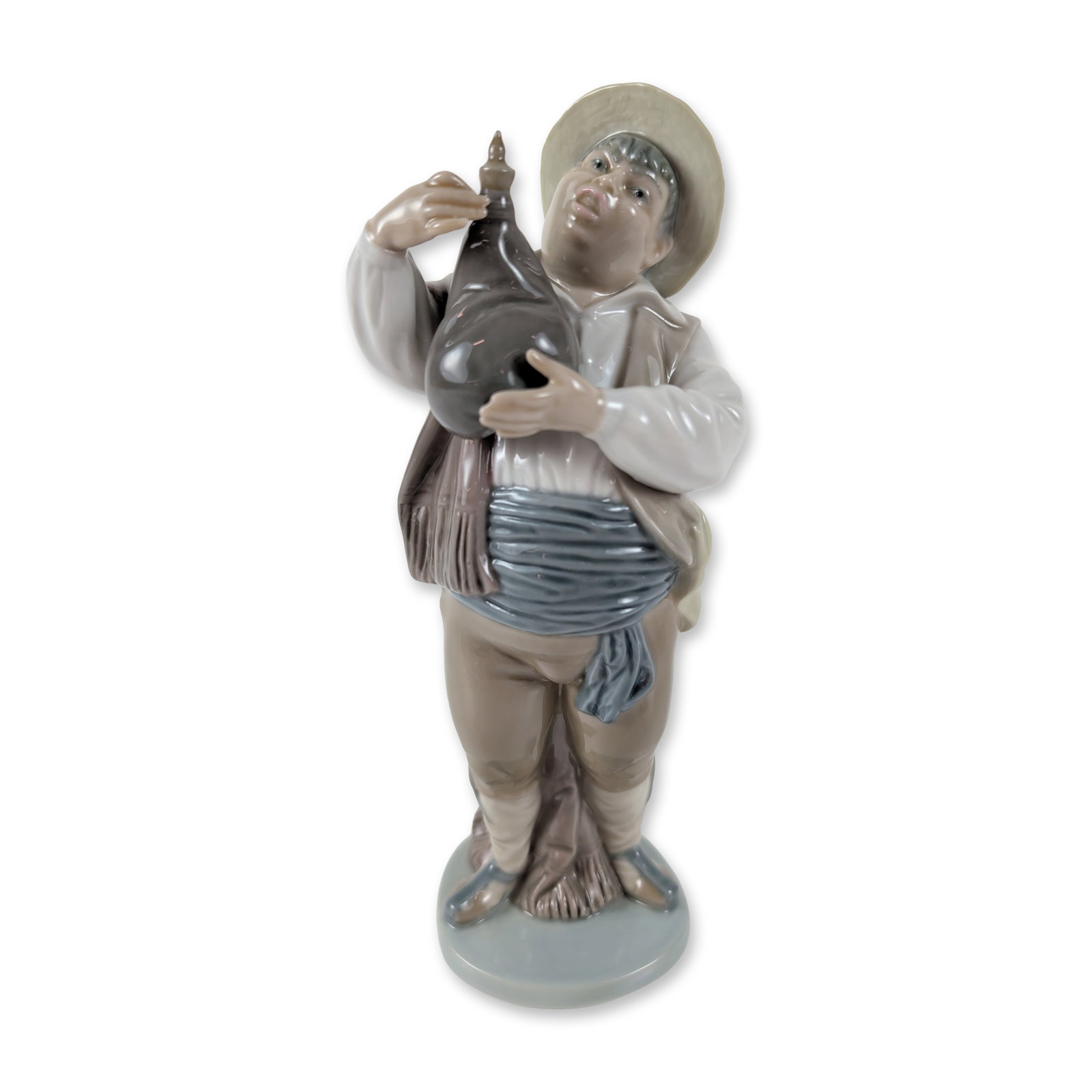 Lladro Porcelain Figurine, A Toast by Sancho 1005165: The figure is depicted in traditional peasant attire, wearing a wide-brimmed tan hat, white long-sleeved shirt, brown trousers, and a blue and white striped waist sash. The figure holds a dark brown w
