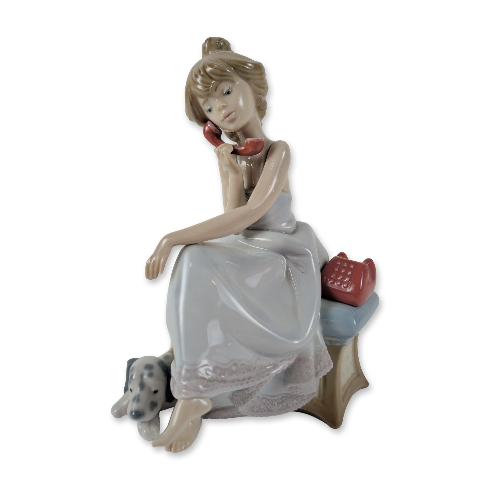Lladro Porcelain Figurine, Chit Chat 1005466: The piece depicts a young girl in a pale blue nightgown seated on a stool, holding a red telephone receiver to her ear. A spotted Dalmatian puppy rests at the base of the stool. The figurine features 