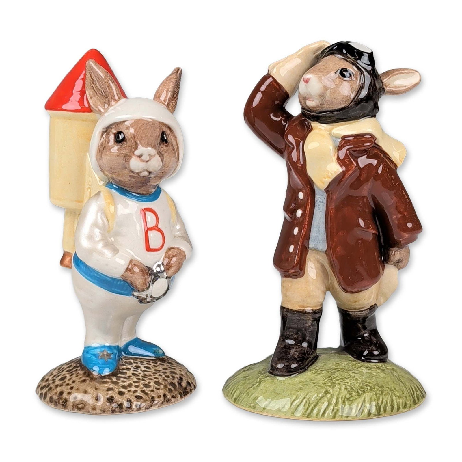 2pc Royal Doulton Bunnykins Rocket Man and Airman Figurines: Royal Doulton. A pair of Bunnykins figurines in glazed ceramic. The grouping includes Astro Bunnykins Rocket Man (DB 20), featuring a rabbit in a white space suit with blue star-motif boots and a red-