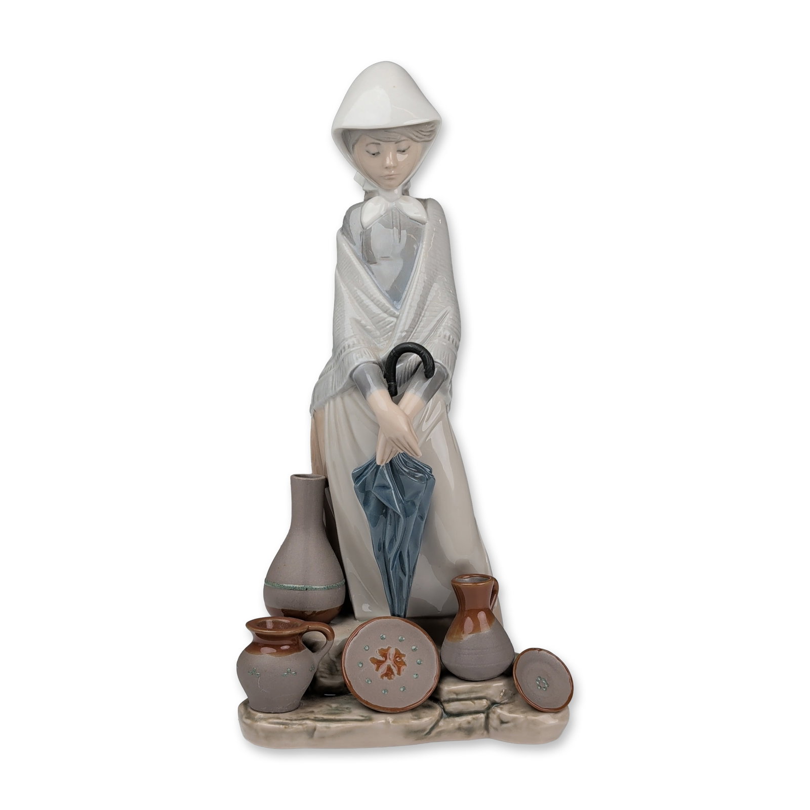 Lladro Porcelain Figurine, Ceramic Seller Woman 01005081: The piece depicts a young woman in traditional attire featuring a white headpiece and a draped dress in grey and tan tones. She is shown standing and holding a folded blue umbrella with a black handle