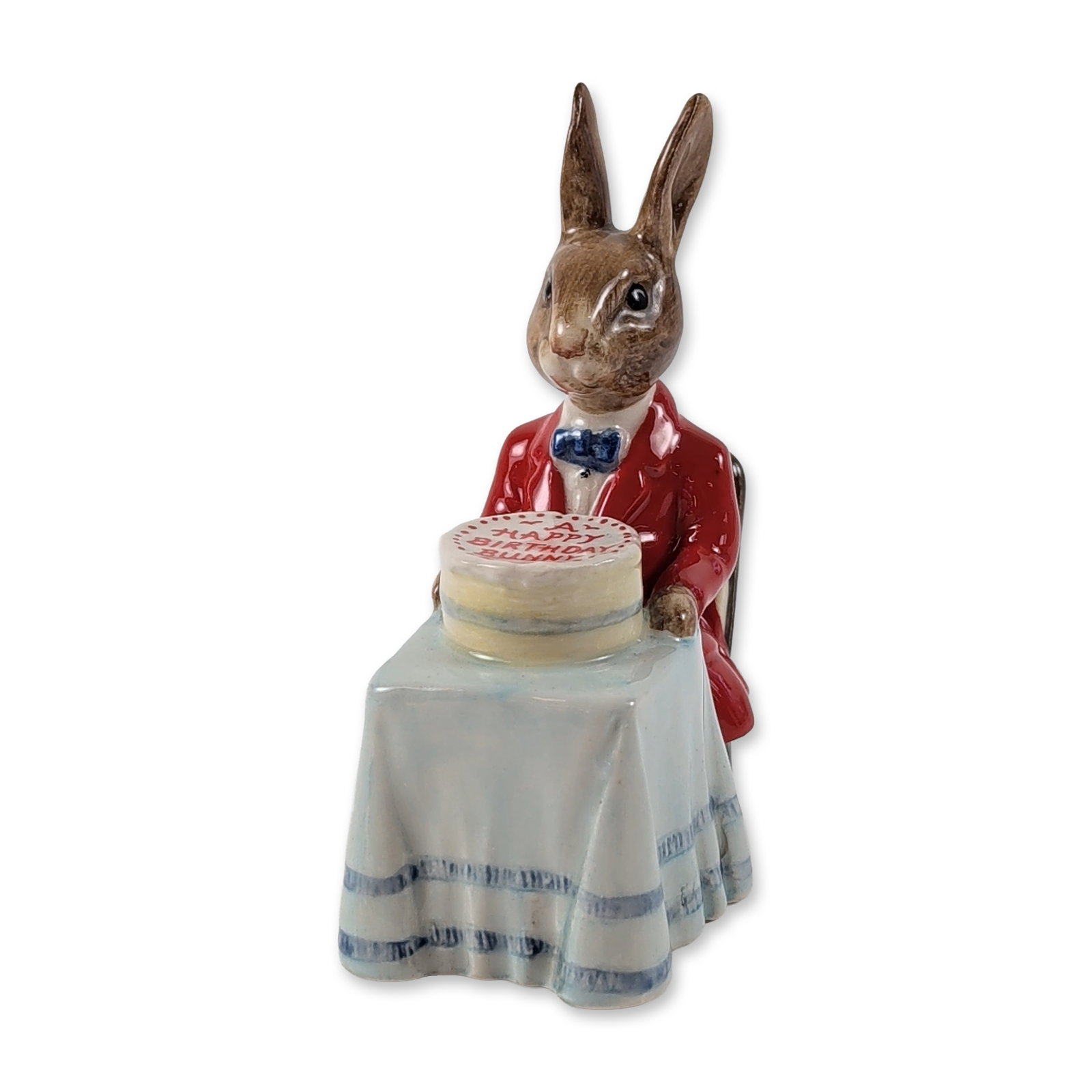 Royal Doulton Happy Birthday Bunnykins Figurine DB21: Royal Doulton. Happy Birthday Bunnykins figurine in polychrome glazed ceramic, model DB21, depicting an anthropomorphic rabbit in a red jacket and blue bow tie seated at a table with a birthday cake. 