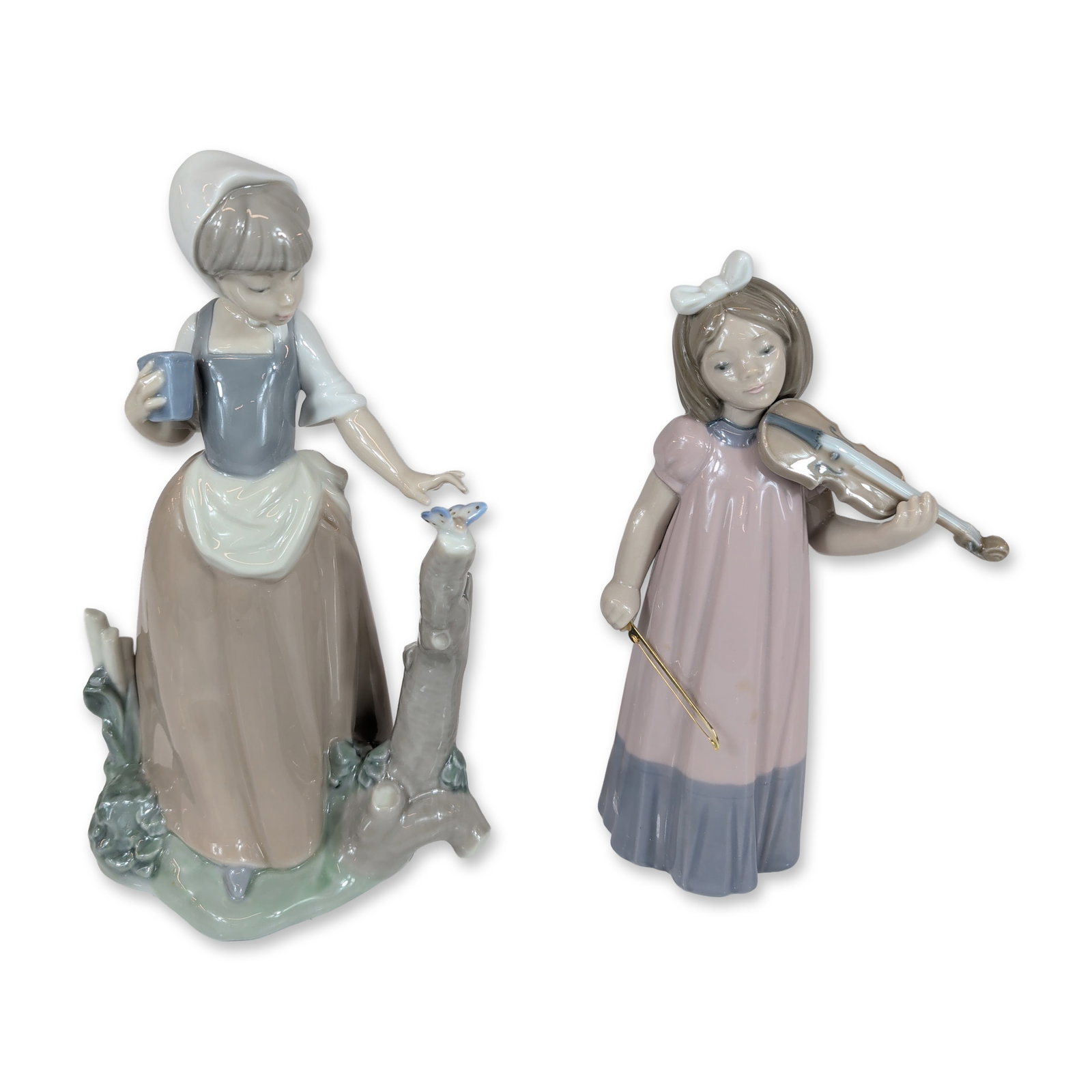 2pc Nao by Lladro Porcelain Figurines: Nao by Lladro. Pair of glazed porcelain figurines featuring childhood subjects. The first depicts a young girl with a white hair bow playing a violin, dressed in a pink gown with a blue-grey border. T