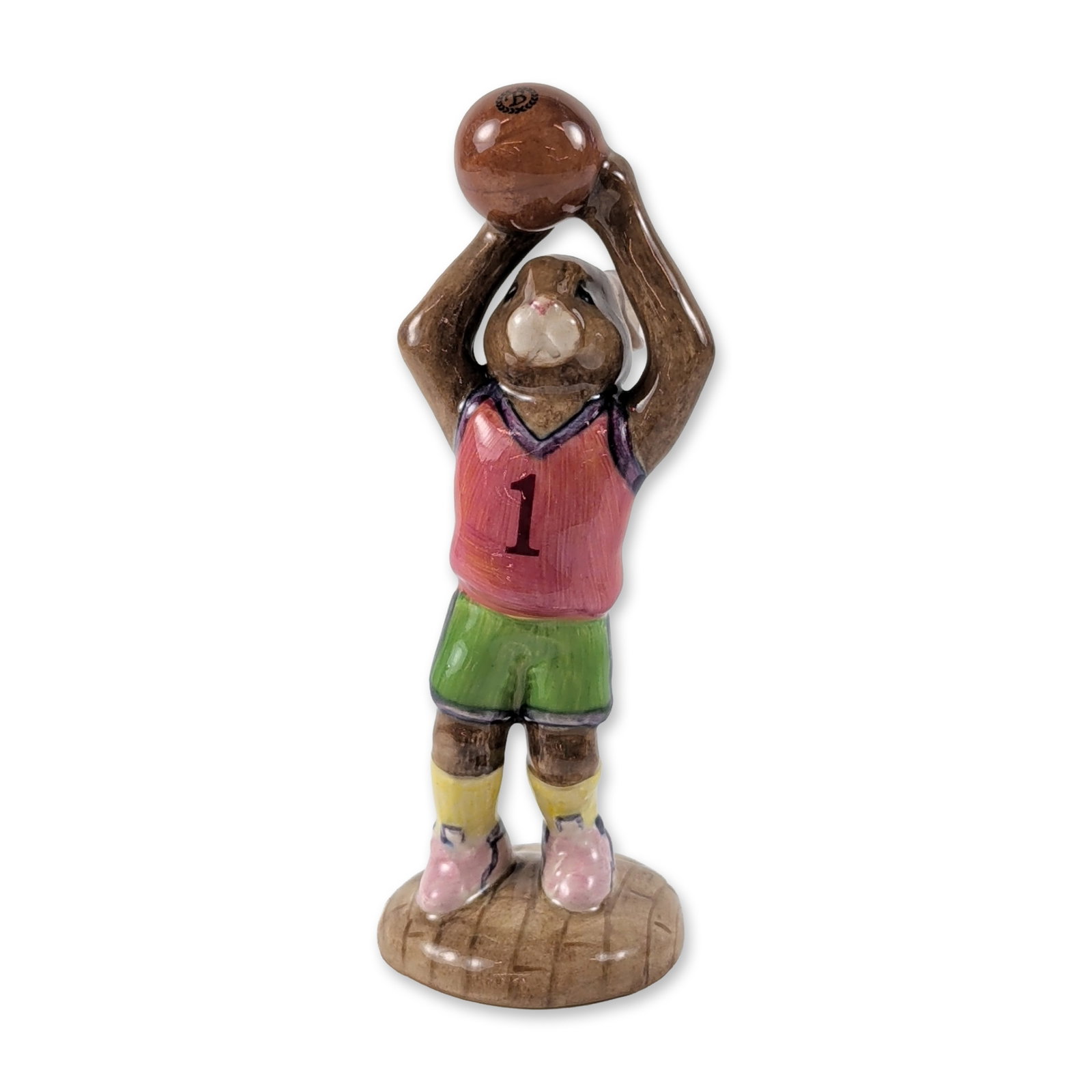 Royal Doulton Basketball Bunnykins Limited Edition Figurine: Royal Doulton. Basketball Bunnykins figurine in hand-decorated ceramic, featuring a brown rabbit in a red, green, and pink basketball uniform on a wood-grain base. The piece is hand-marked number 986 